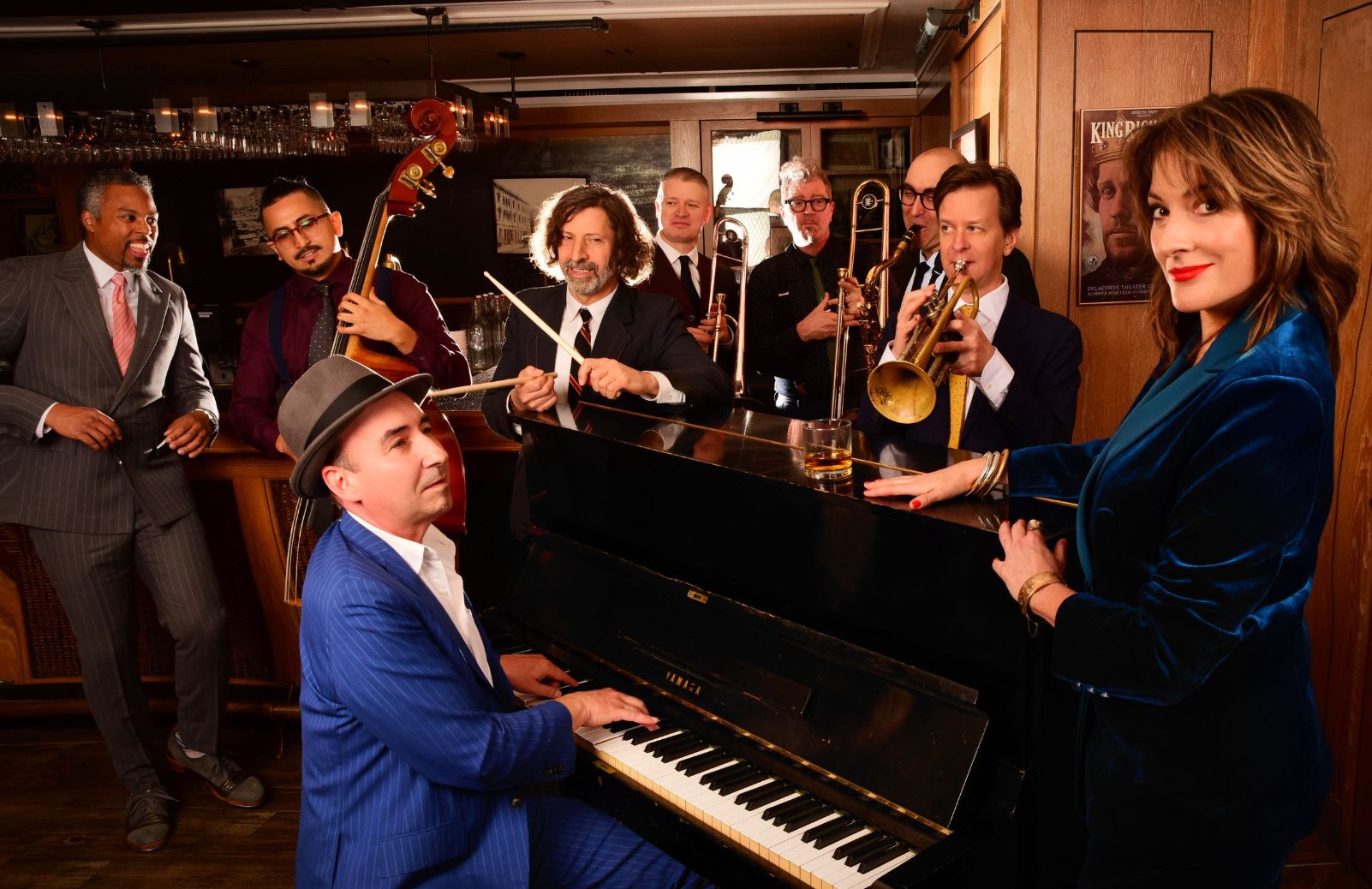 A jazz ensemble gathers around an upright piano in a warmly lit bar, with brass, bass, and a vocalist in a teal dress.