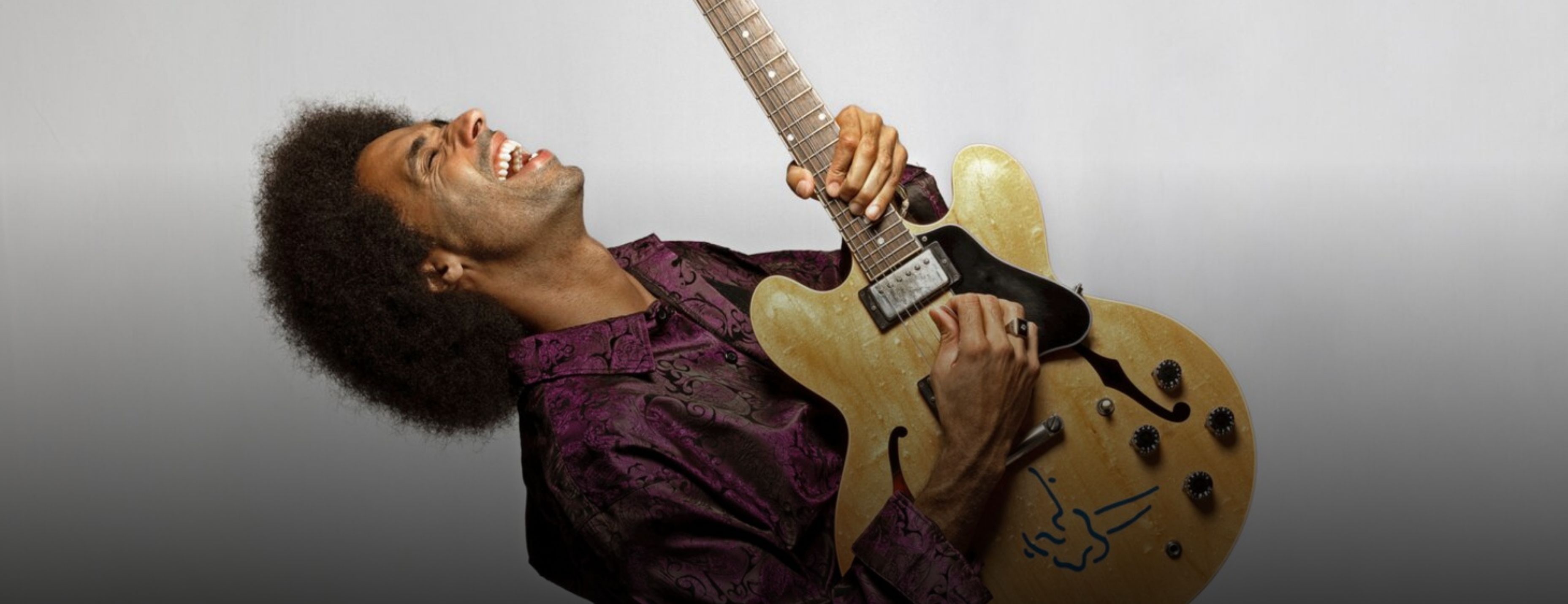 A guitarist throws his head back laughing while playing an electric guitar, wearing a purple patterned shirt against a grey background.
