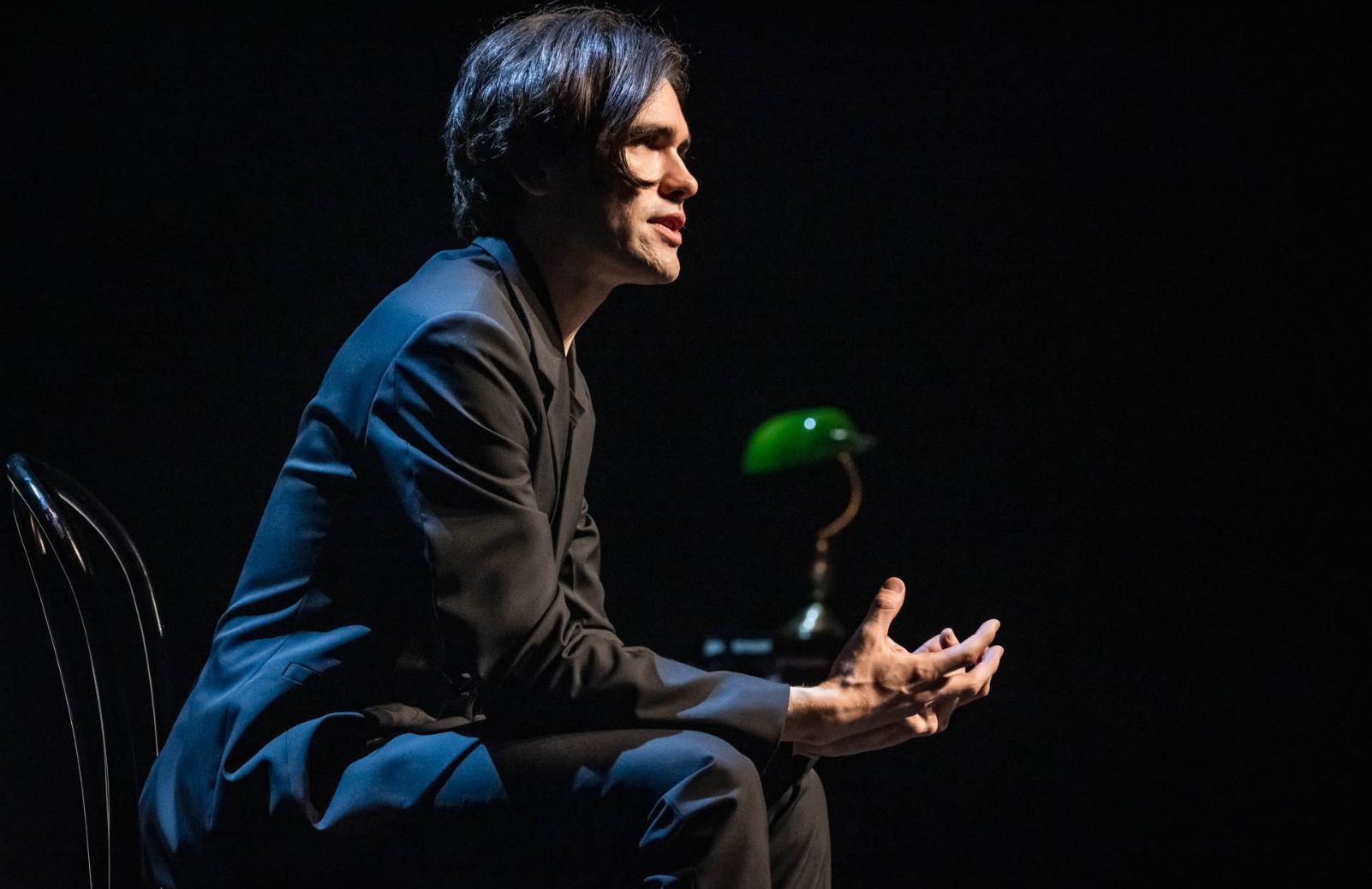 A performer in a black suit sits and gestures on a darkened stage, a green desk lamp glowing behind him.