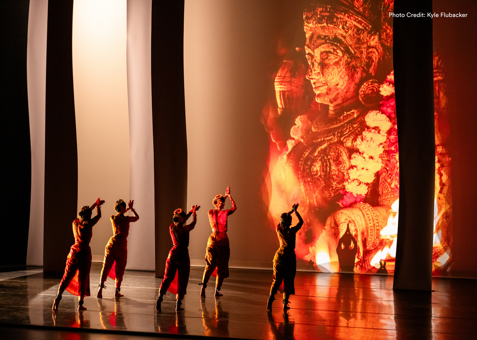 Five classical Indian dancers face away from the camera with arms raised in unison, silhouetted against a massive projected image of a deity engulfed in flame. Photo: Kyle Flubacker.