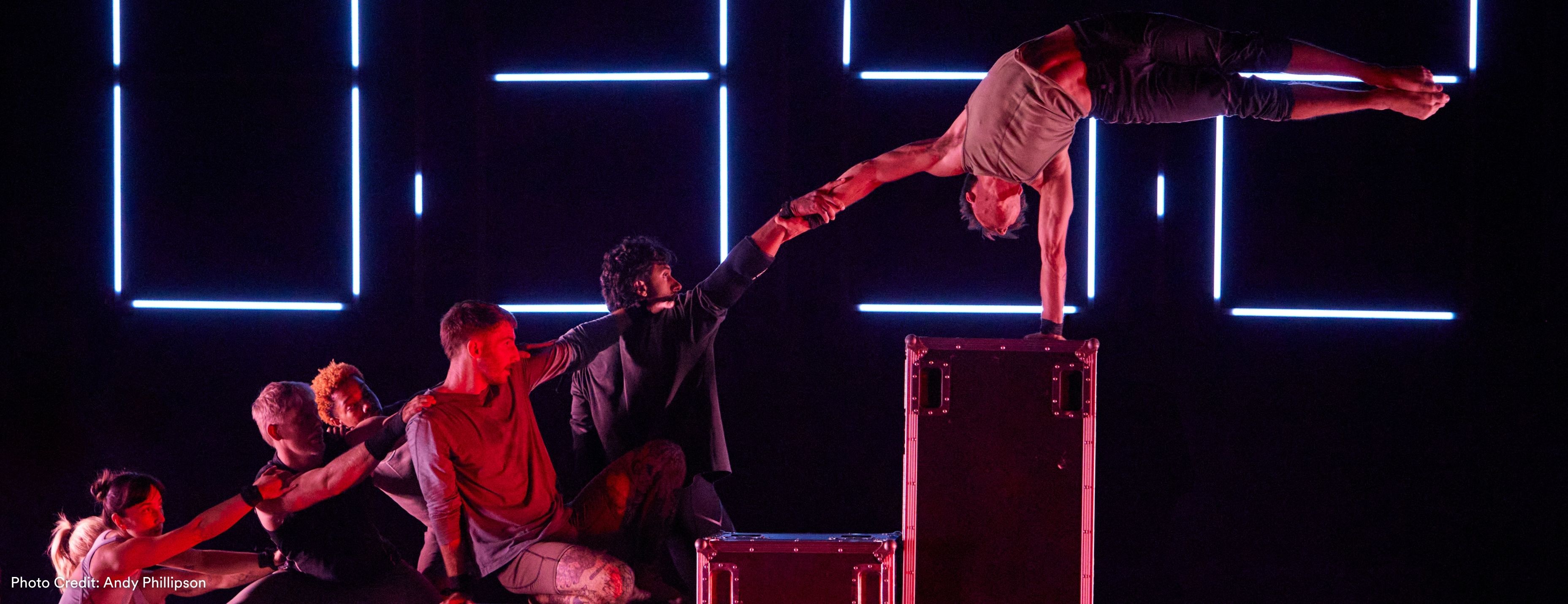 A performer balances horizontally atop a road case, held by a line of other performers' outstretched arms, against a neon digital clock backdrop on a darkened red-lit stage.