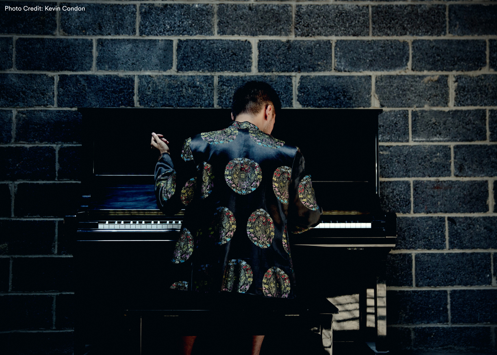 A pianist seen from behind at an upright piano against a dark brick wall, wearing a bold black jacket with large circular embroidered medallions. Photo: Kevin Condon.