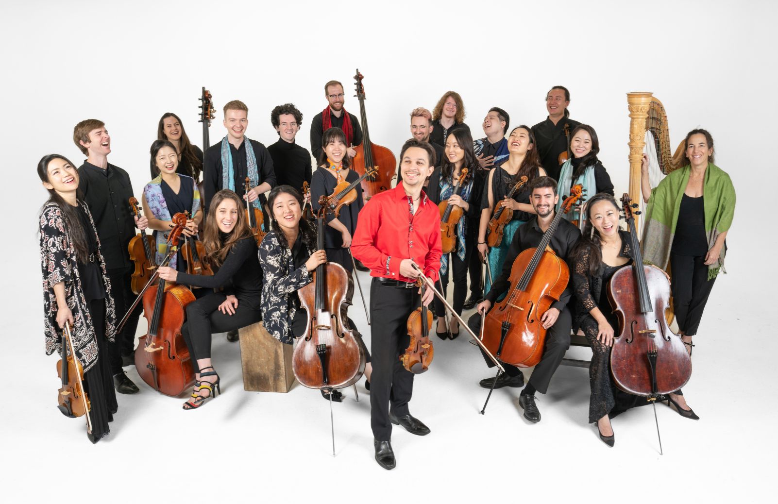 An ensemble poses with their instruments against a white background, led by a conductor in a red shirt at center.