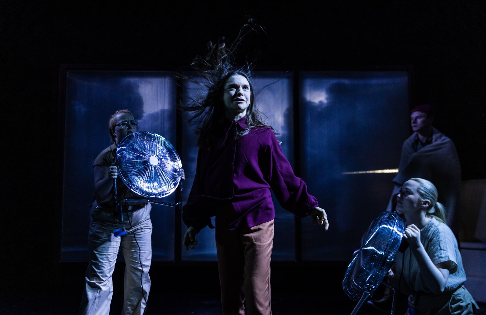 A performer's hair whips in the wind on a dark stage as cast members hold electric fans around her.