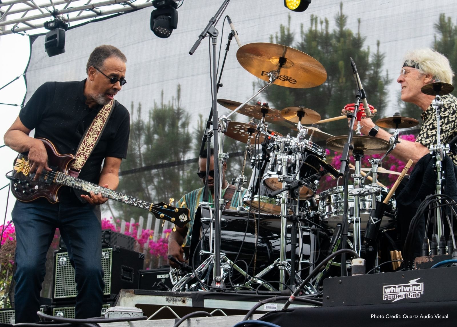Two musicians, a bassist and a drummer, perform energetically on an outdoor stage framed by pink bougainvillea.