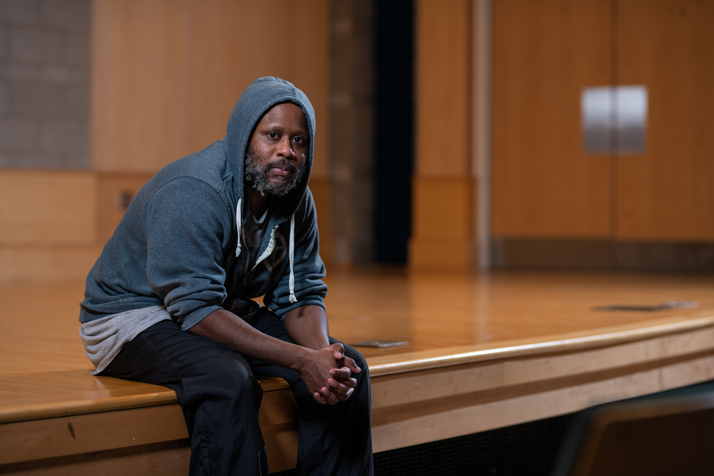 Shodekeh Talifero sits on the edge of a wooden stage with the hood of his sweatshirt on, hands clasped together in his lap.
