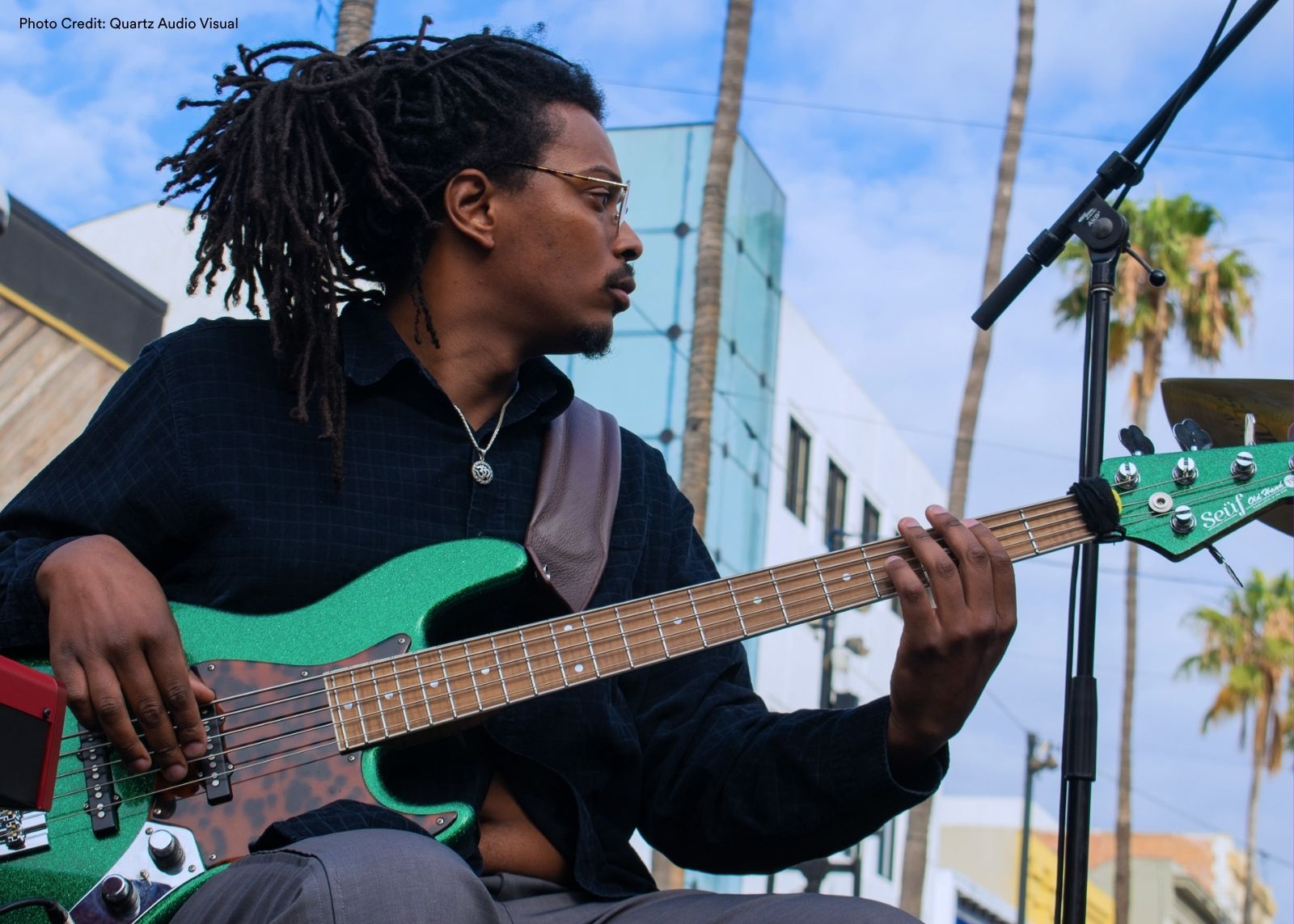 A bassist with gold-rimmed glasses plays a green electric bass guitar outdoors, palm trees visible against a blue sky in the background.