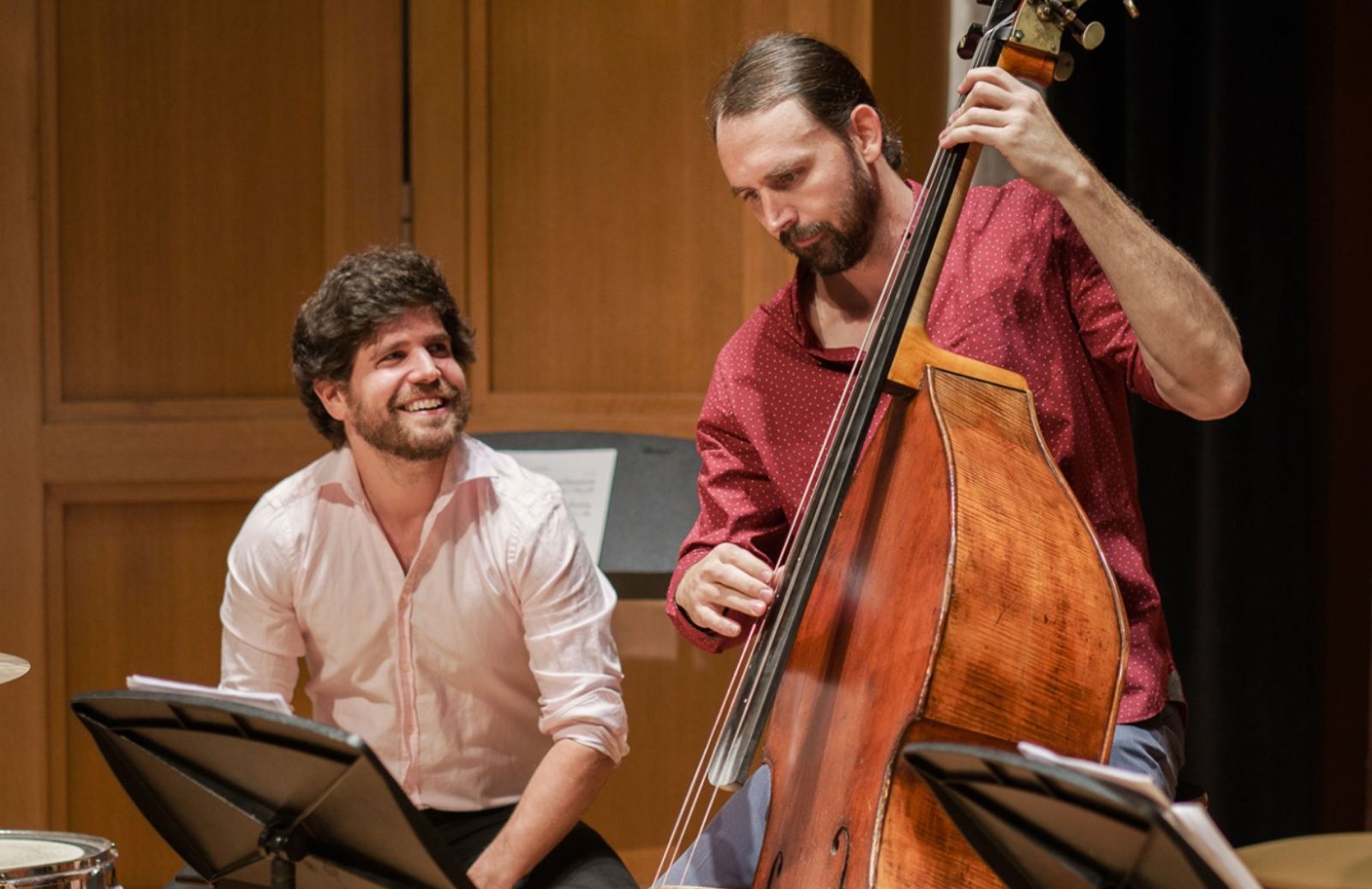 Two musicians performing in front of a wooden wall, one with a cello and the other with drums.