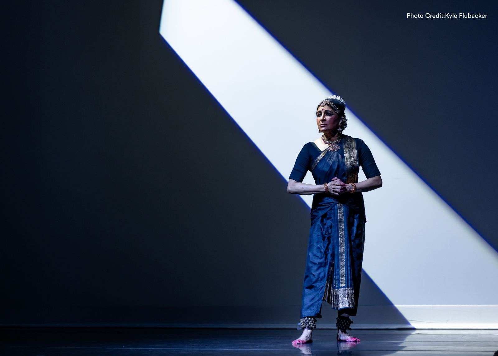 A soloist in a navy sari stands in a beam of dramatic diagonal light on an otherwise dark stage. Photo: Kyle Flubacker.