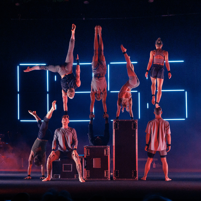 Acrobatic performers hold handstands on cases and each other against glowing neon clock digits on a dramatic stage. 