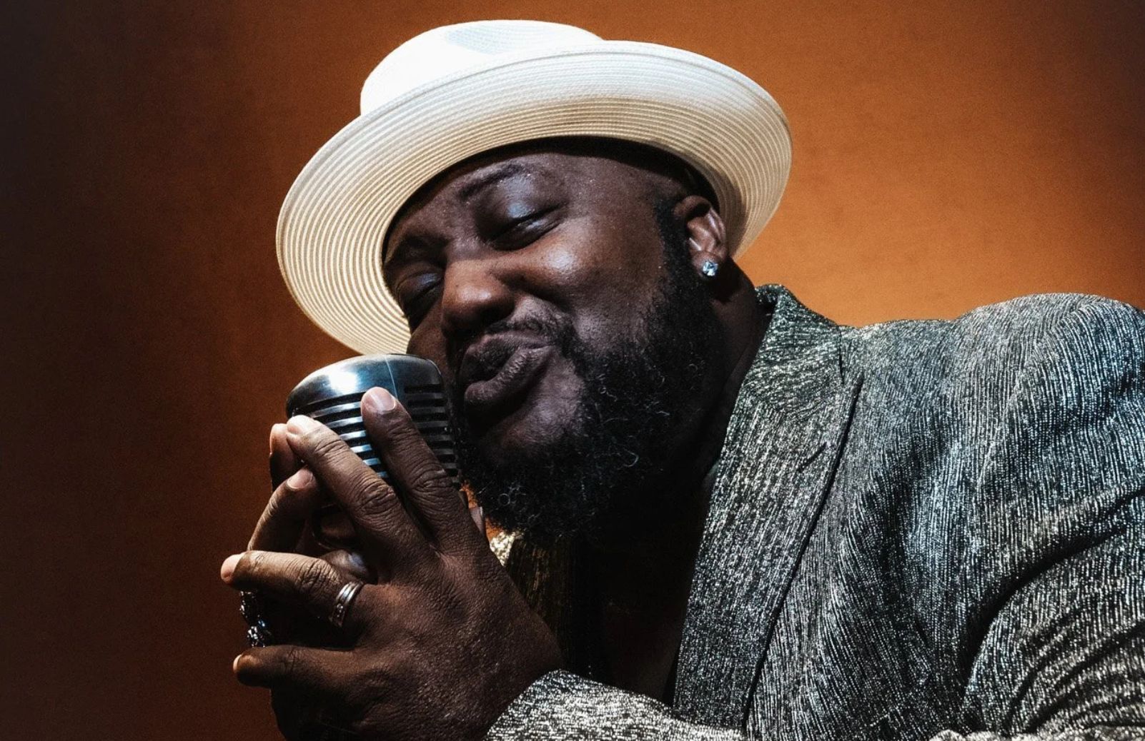 A singer in a white hat and gray jacket performs with eyes closed, holding a vintage-style microphone against a warm amber background.