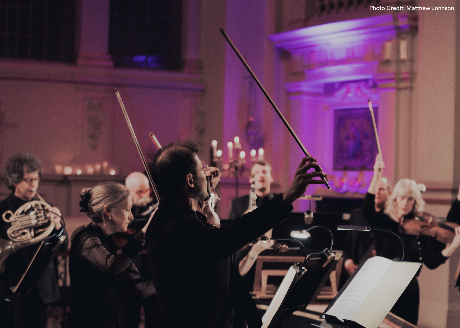 An ensemble in a candlelit church bathed in purple light, with violinists and a French horn player visible around him. Photo: Matthew Johnson.