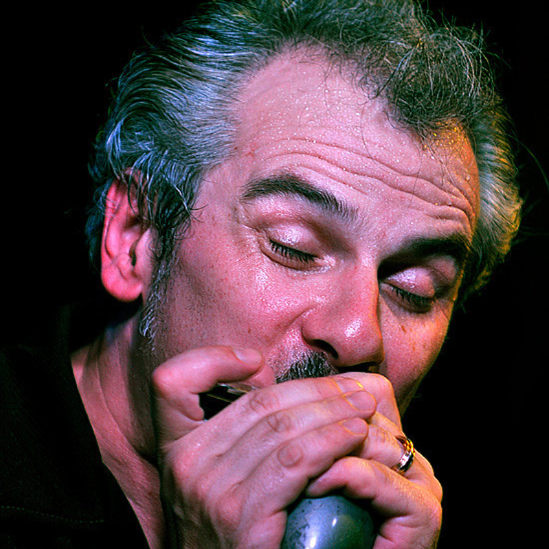 A man with white hair plays the harmonica in front of a black background.