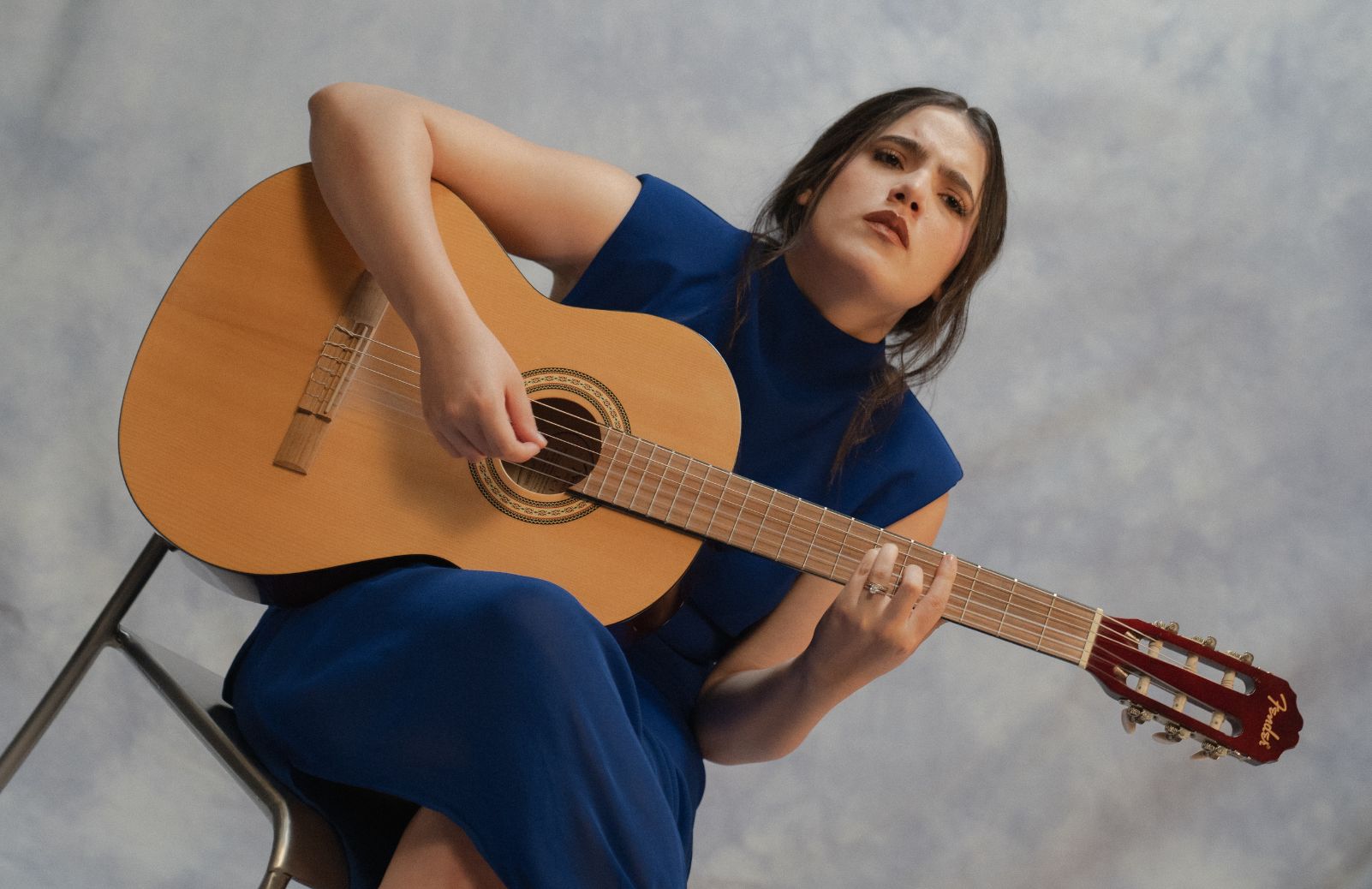 A woman in a blue dress holds a classical guitar while seated.