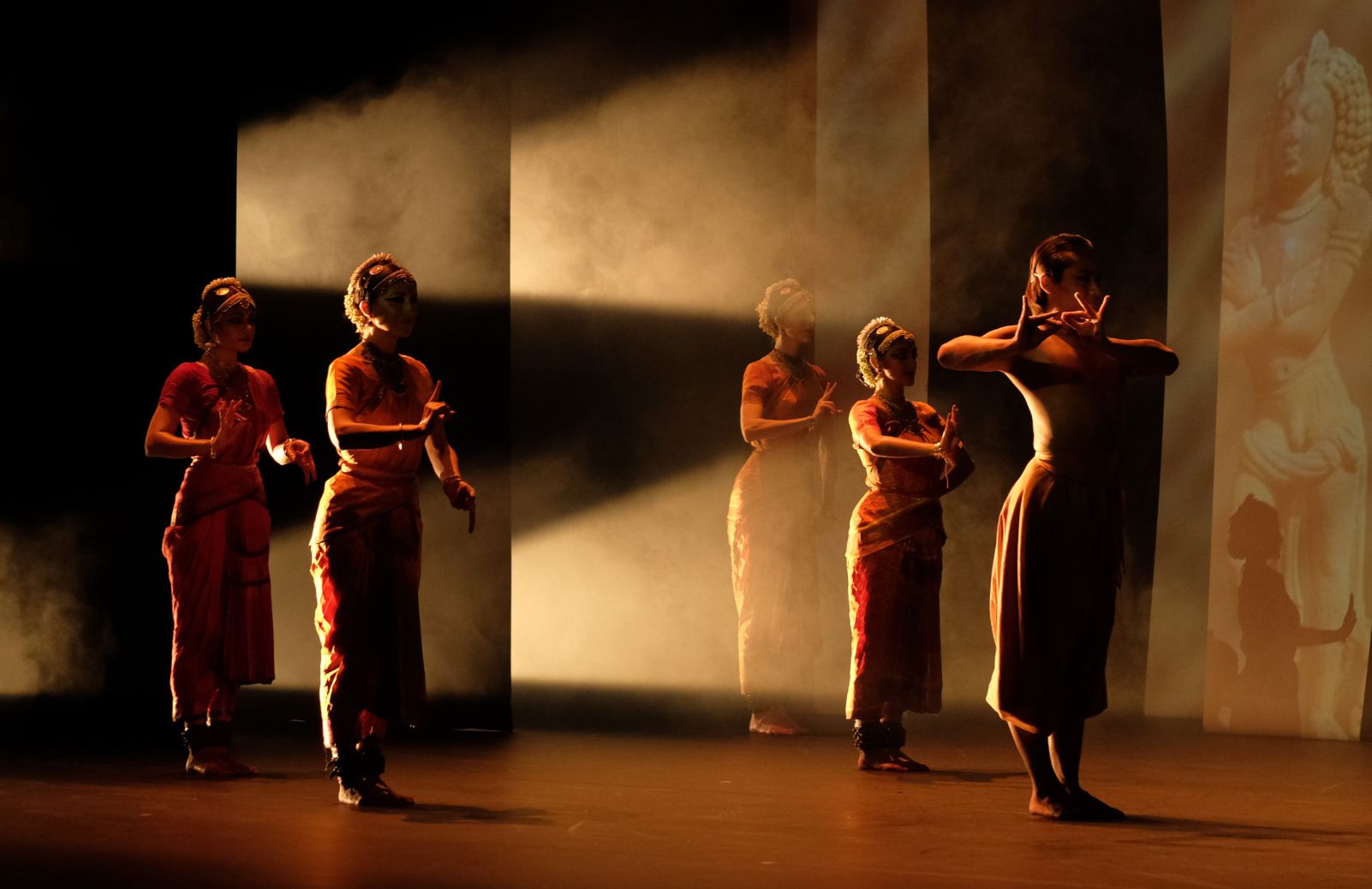 Five dancers in red and gold costumes perform classical Indian dance on a warmly lit stage with projected imagery in the background.