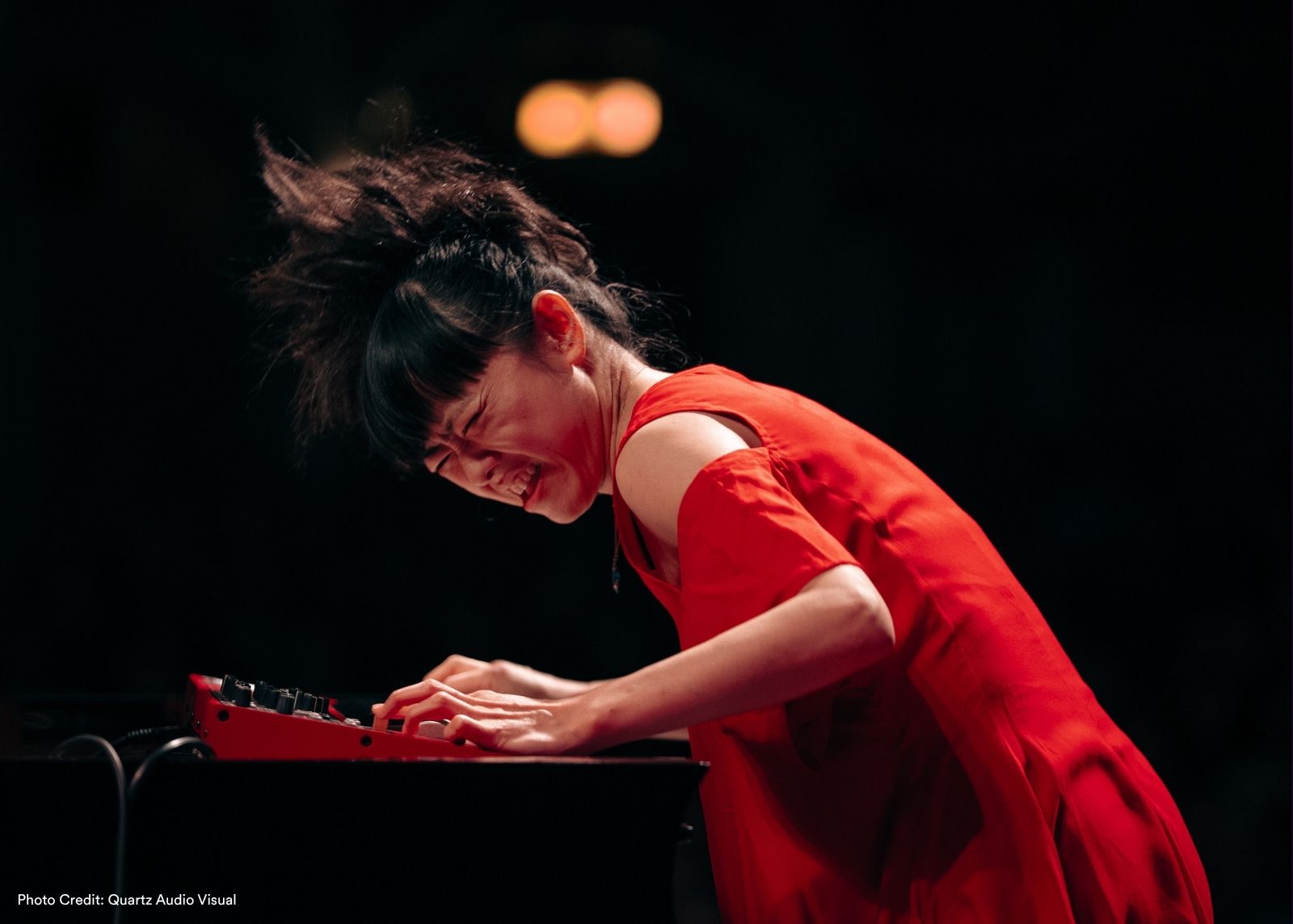 A performer in a red dress leans over a red keyboard on a dark stage.