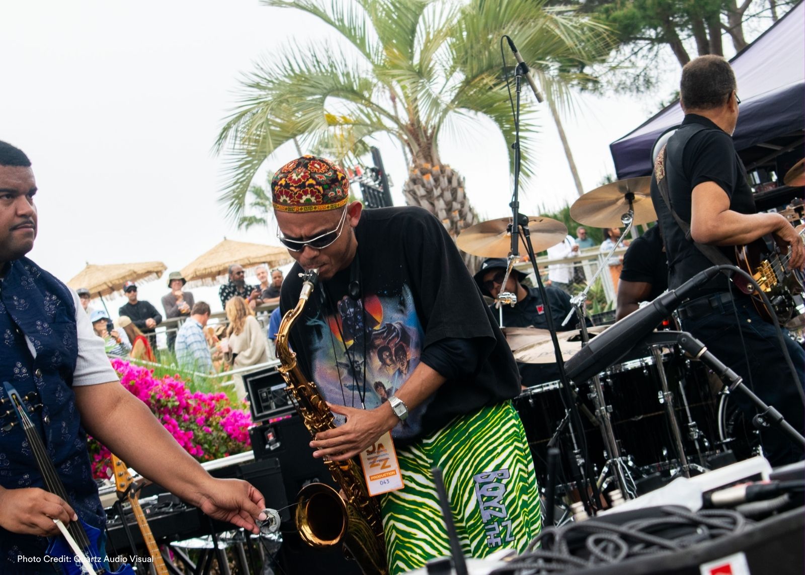 A saxophonist in green zebra-print pants and a colorful kufi plays saxophone on a festival stage, surrounded by bandmates.