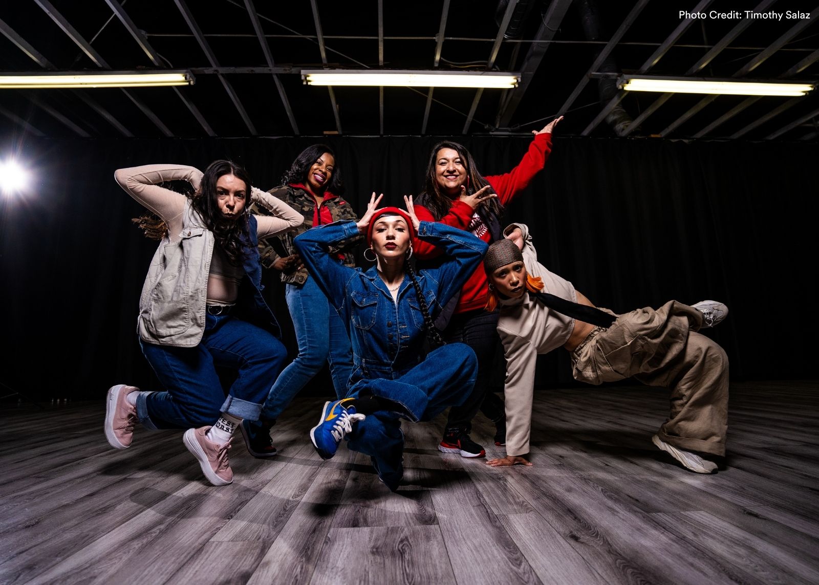 Five women from Versa-Style Dance Company pose together in a studio, with one crouched center in a denim jumpsuit and red beanie as others strike expressive stances around her. Photo: Timothy Salaz.