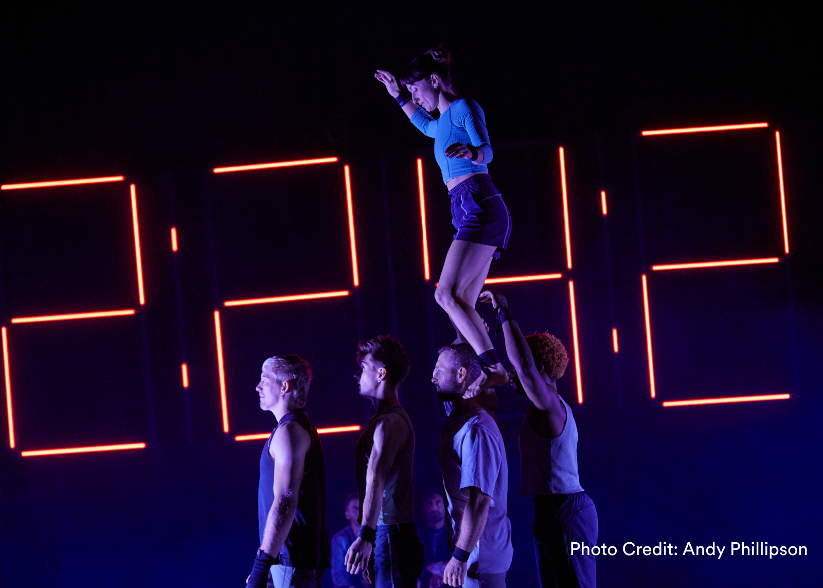 Three acrobats form a tilted diagonal tower on a blue-lit stage as other performers and a live band look on. (Photo Credit: Milissa Martin)