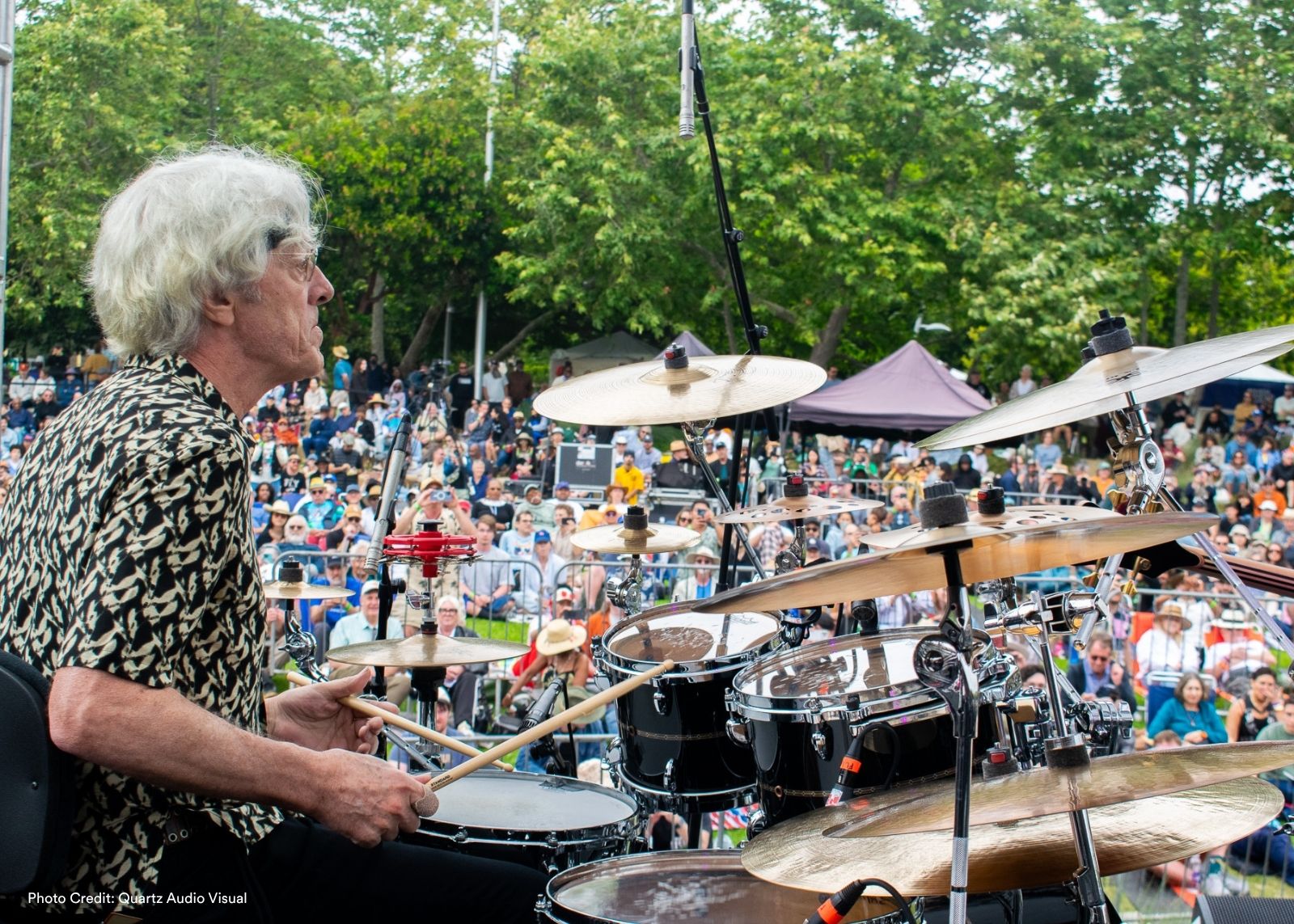 A drummer with white hair performs at an outdoor festival, looking out at a large seated crowd surrounded by trees.