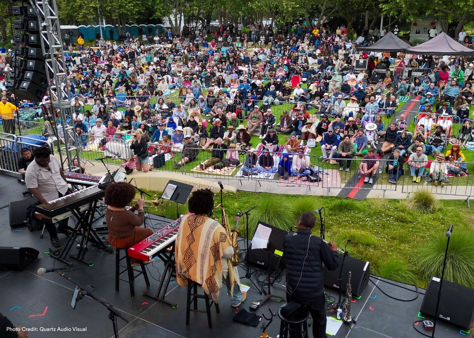 An aerial view of a band performing on an outdoor stage to a massive crowd stretched across a grassy park.