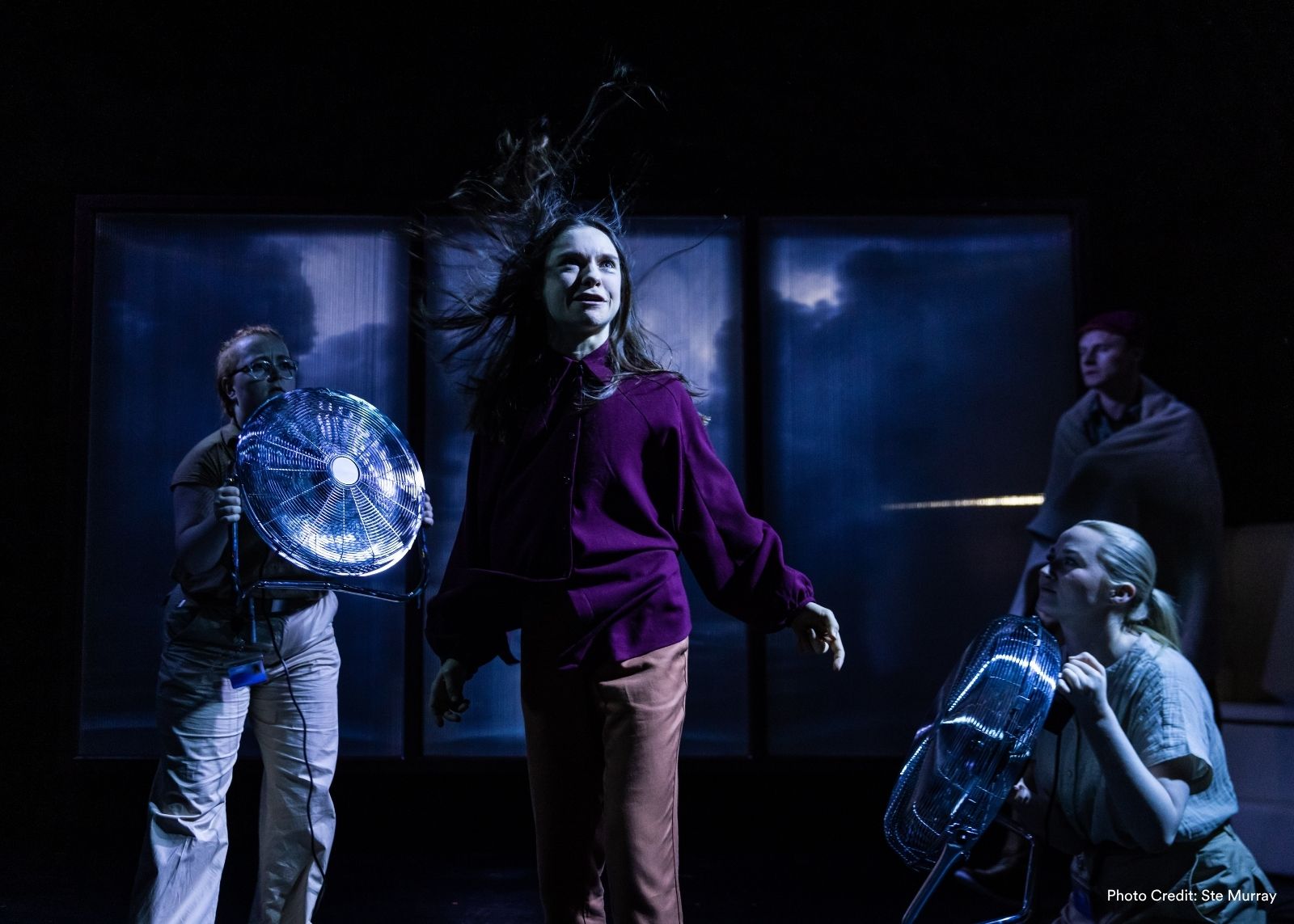 A performer's hair whips upward as cast members aim electric fans at her on a stormy, blue-lit stage.