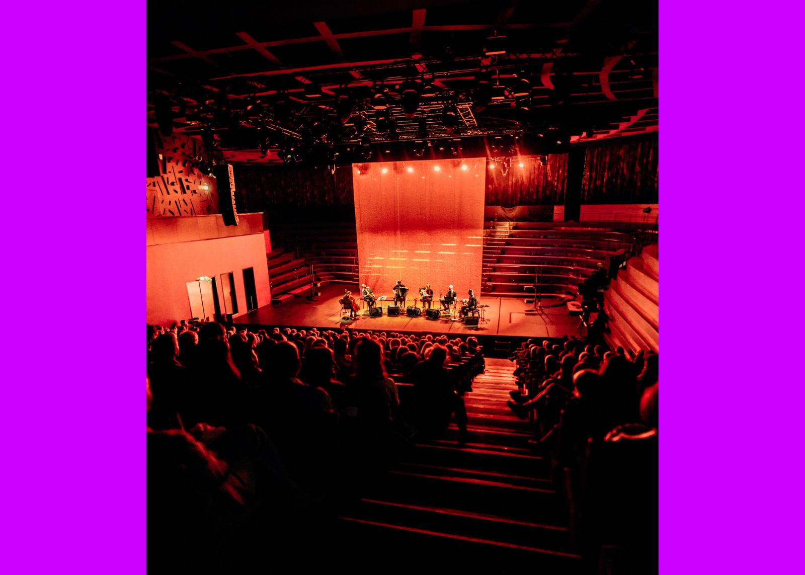 A small ensemble performs on a warmly lit stage in a full concert hall bathed in deep red light.