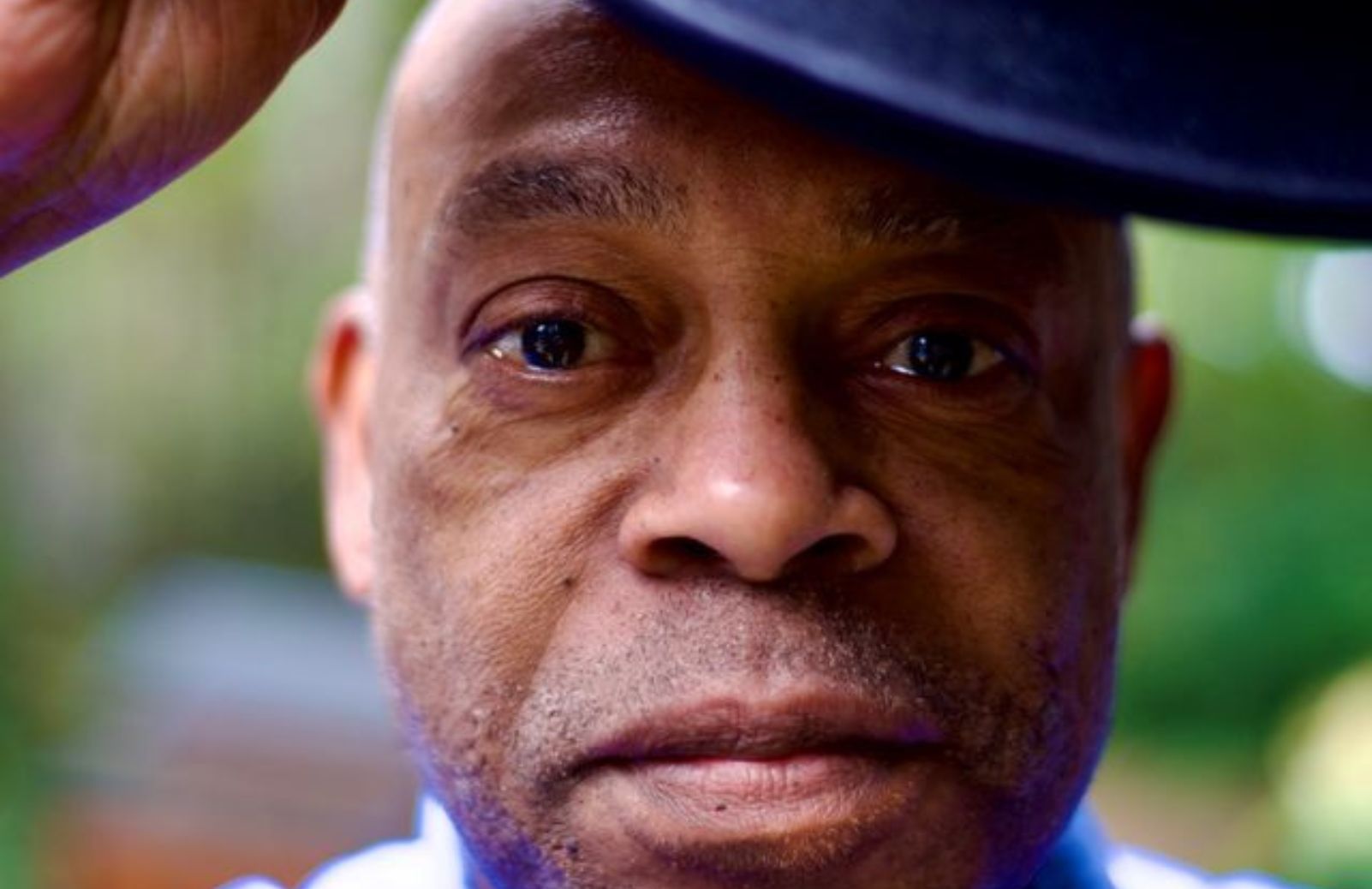 A man tipping his hat toward the camera, with a soft outdoor background.