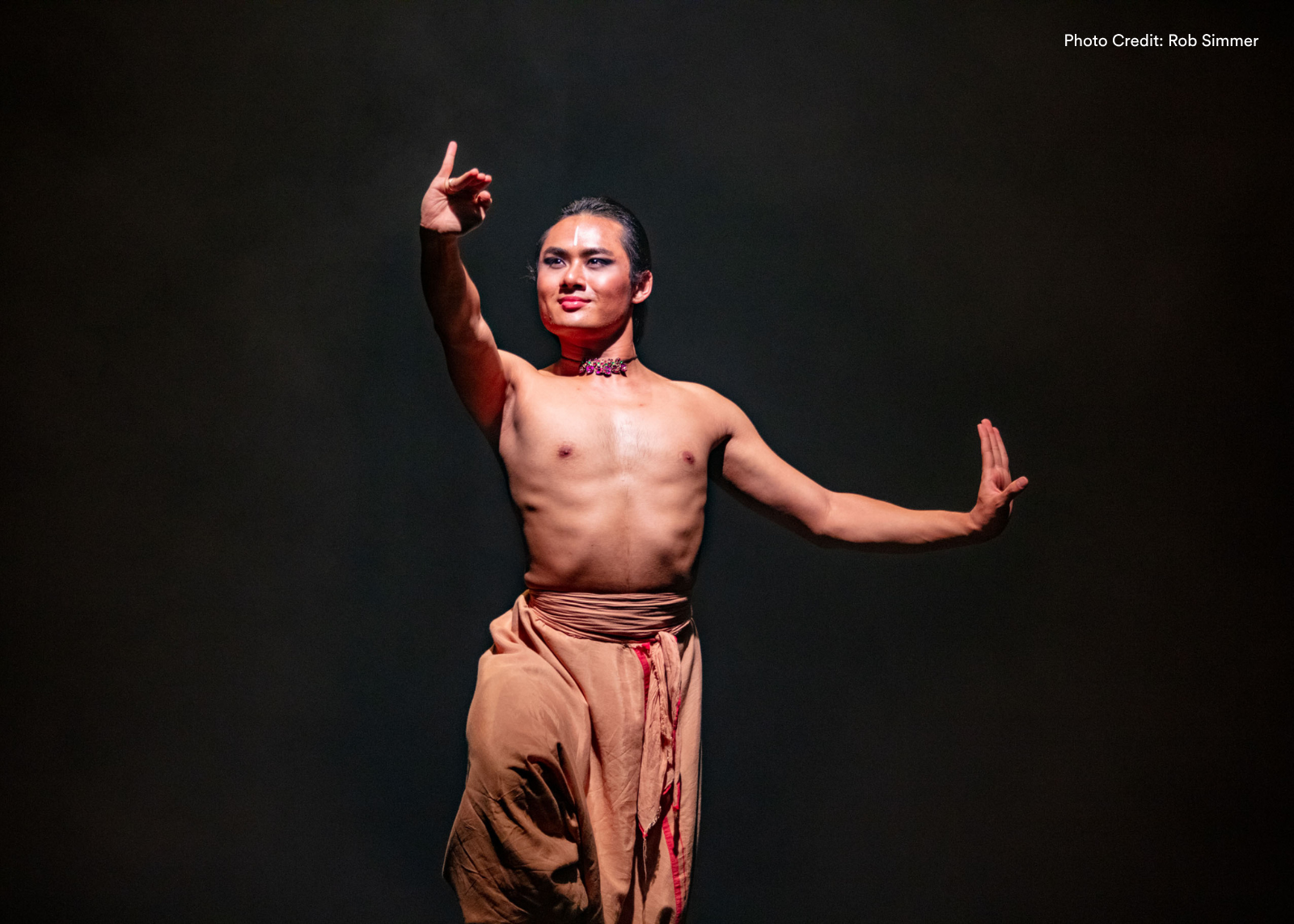 A dancer in tan pants and jewelry performs classical Indian dance against a dark backdrop. Photo: Rob Simmer.