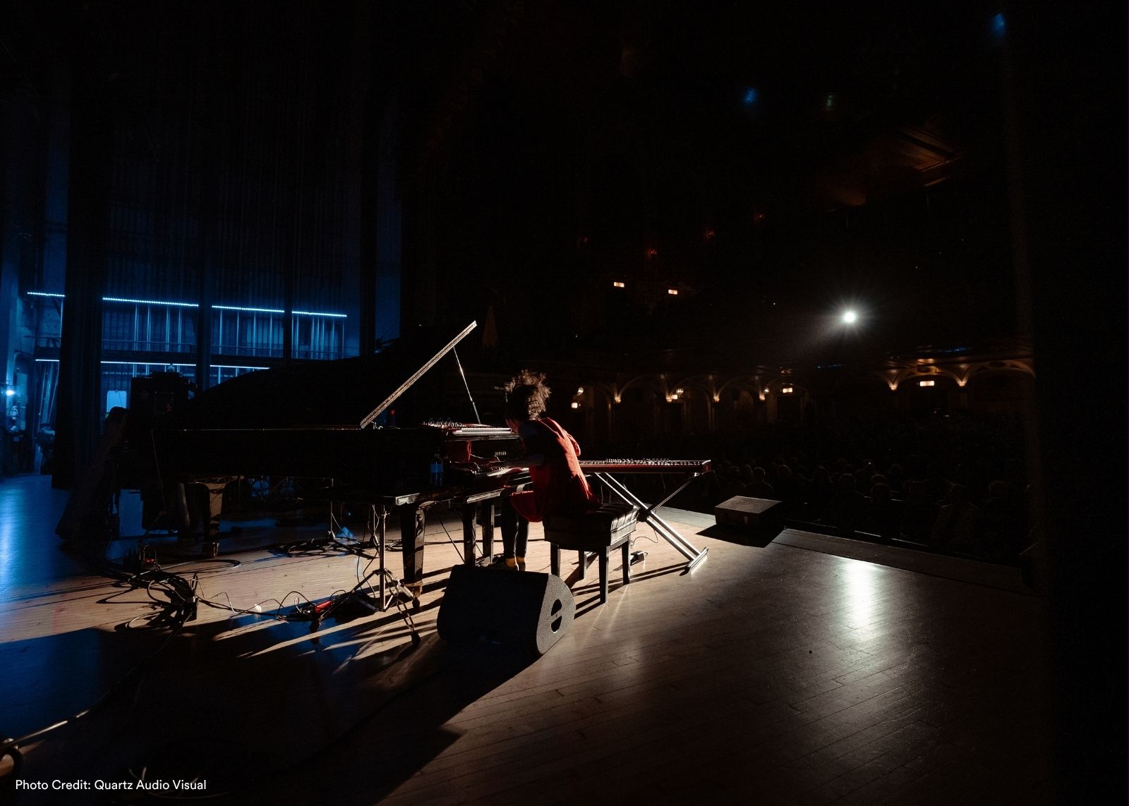 A lone performer in red sits at a grand piano and keyboard rig on a darkened stage, silhouetted against cool blue lighting, facing an audience in a large hall.