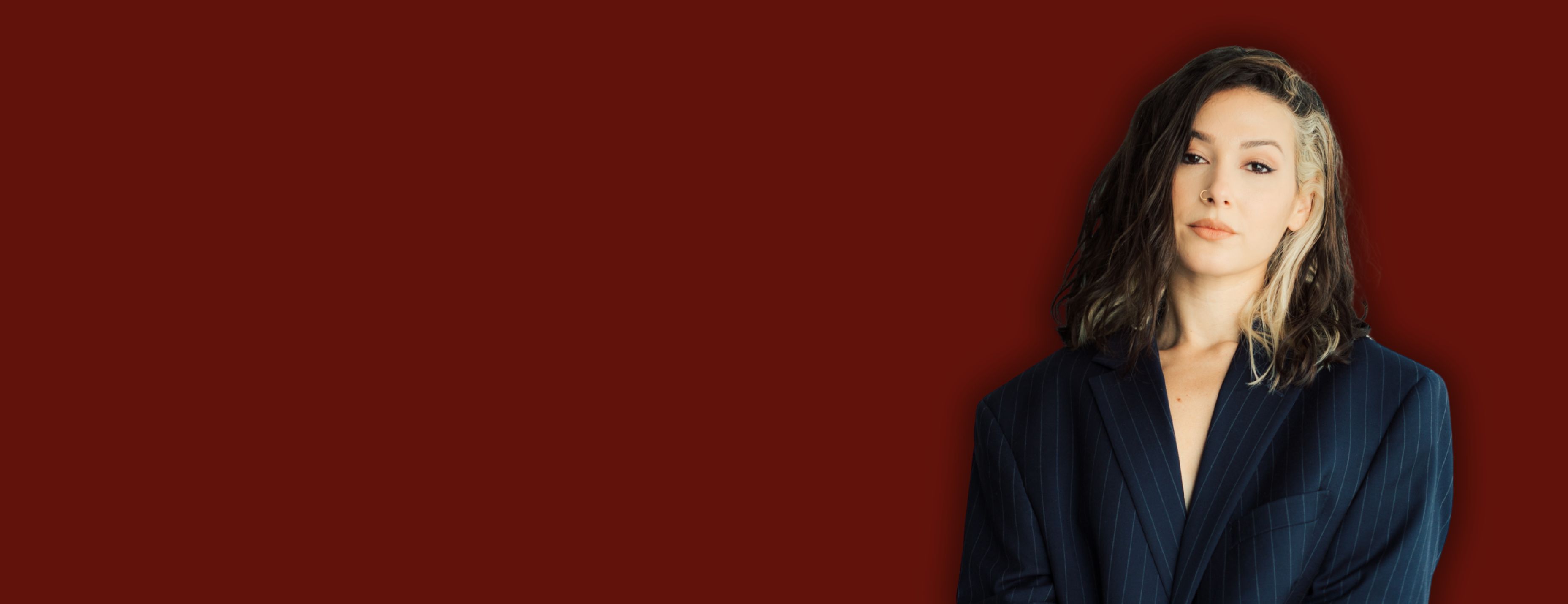 A woman with dark hair gazes at the camera, wearing a navy pinstripe blazer against a deep red background.