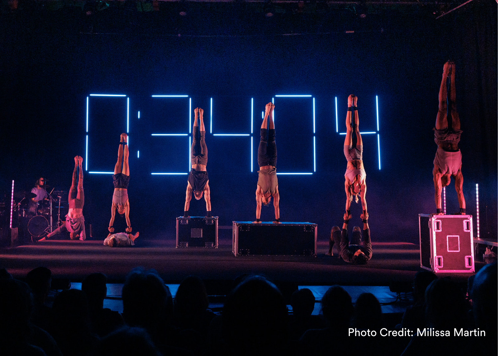 A row of acrobats perform simultaneous handstands across a stage, backed by a large neon countdown clock. (Photo Credit: Milissa Martin)