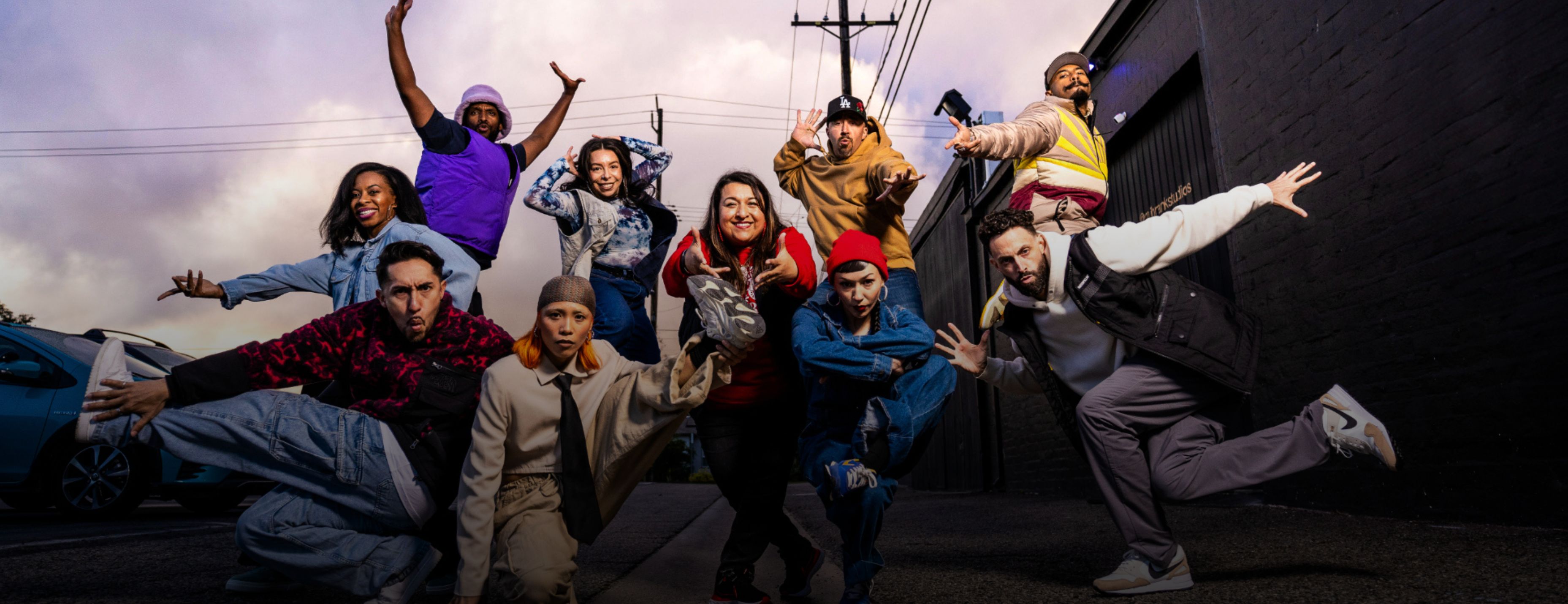 A dance ensemble strikes dynamic, energetic poses together on a city street at dusk, dressed in colorful everyday clothing against a moody purple sky.