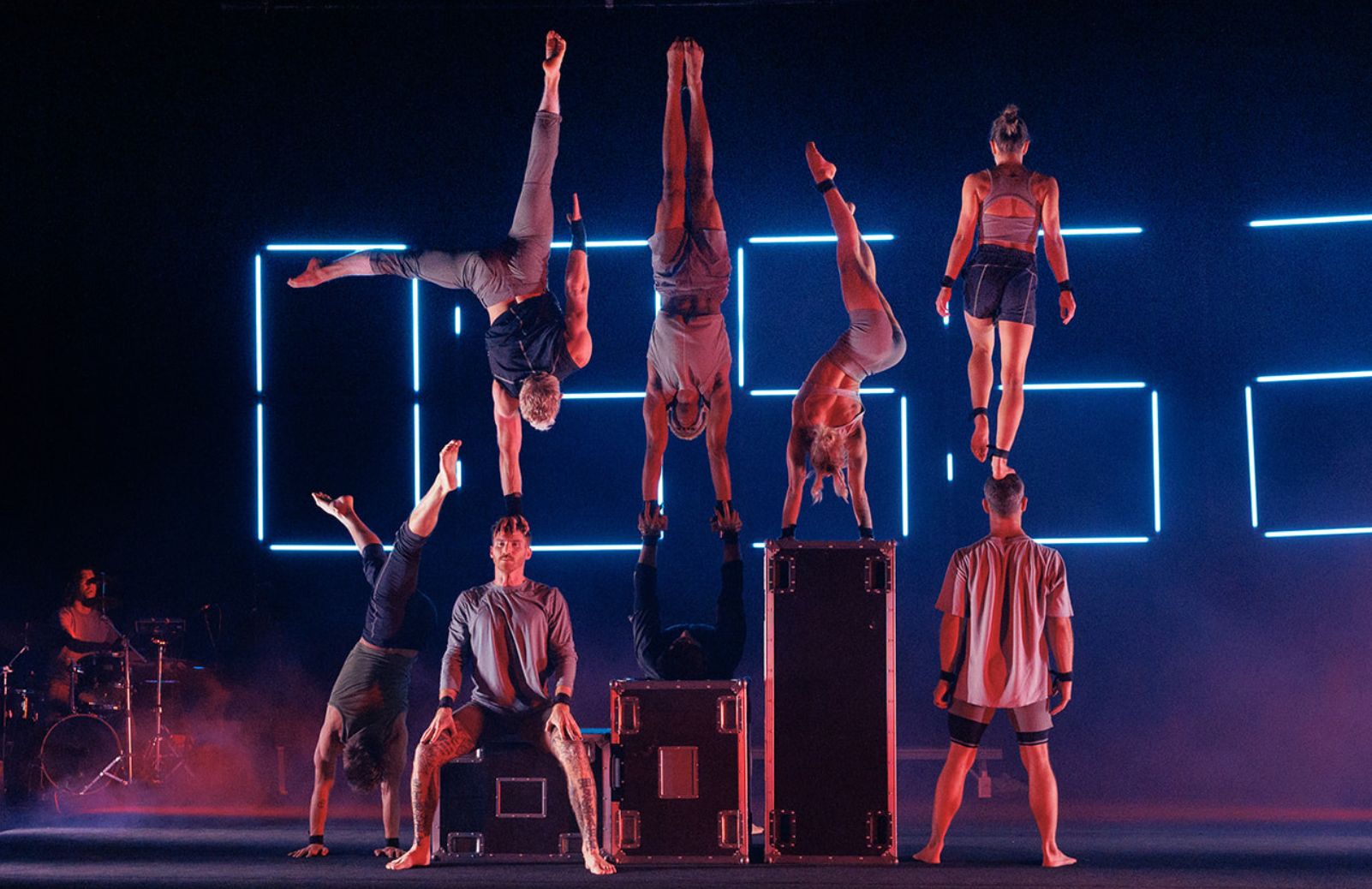 Acrobatic performers hold handstands and balances on a dramatic stage lit by glowing neon clock digits.