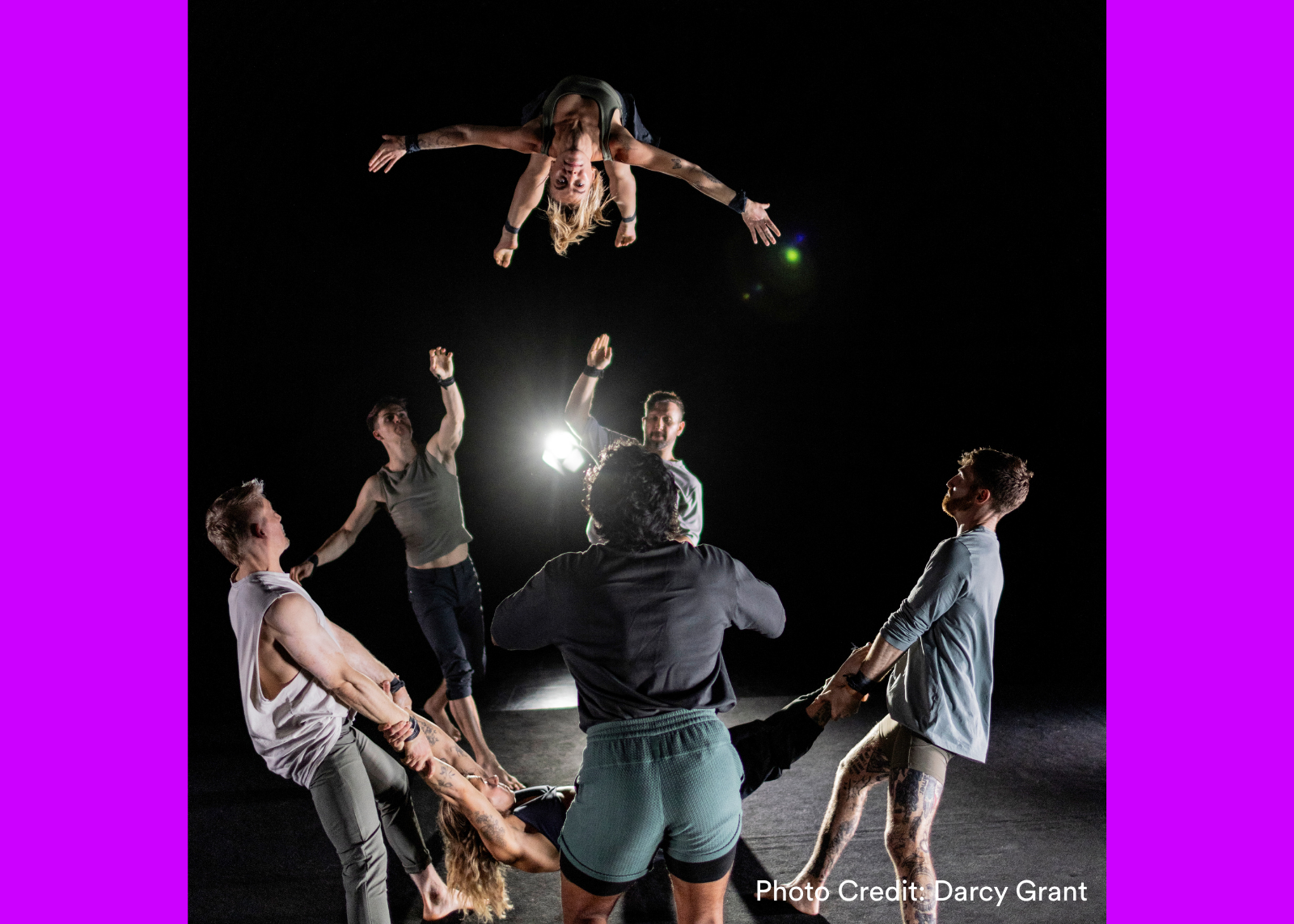 An acrobat is tossed and caught upside down above a circle of performers on a darkened stage. (Photo Credit: Darcy Grant)