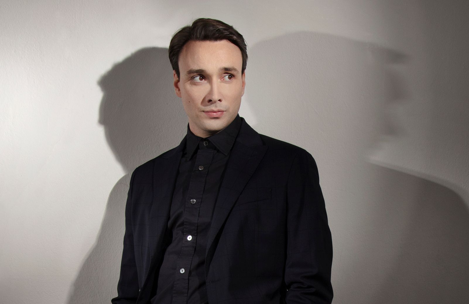 A man in a black suit stands in front of a white background, flanked by shadows.