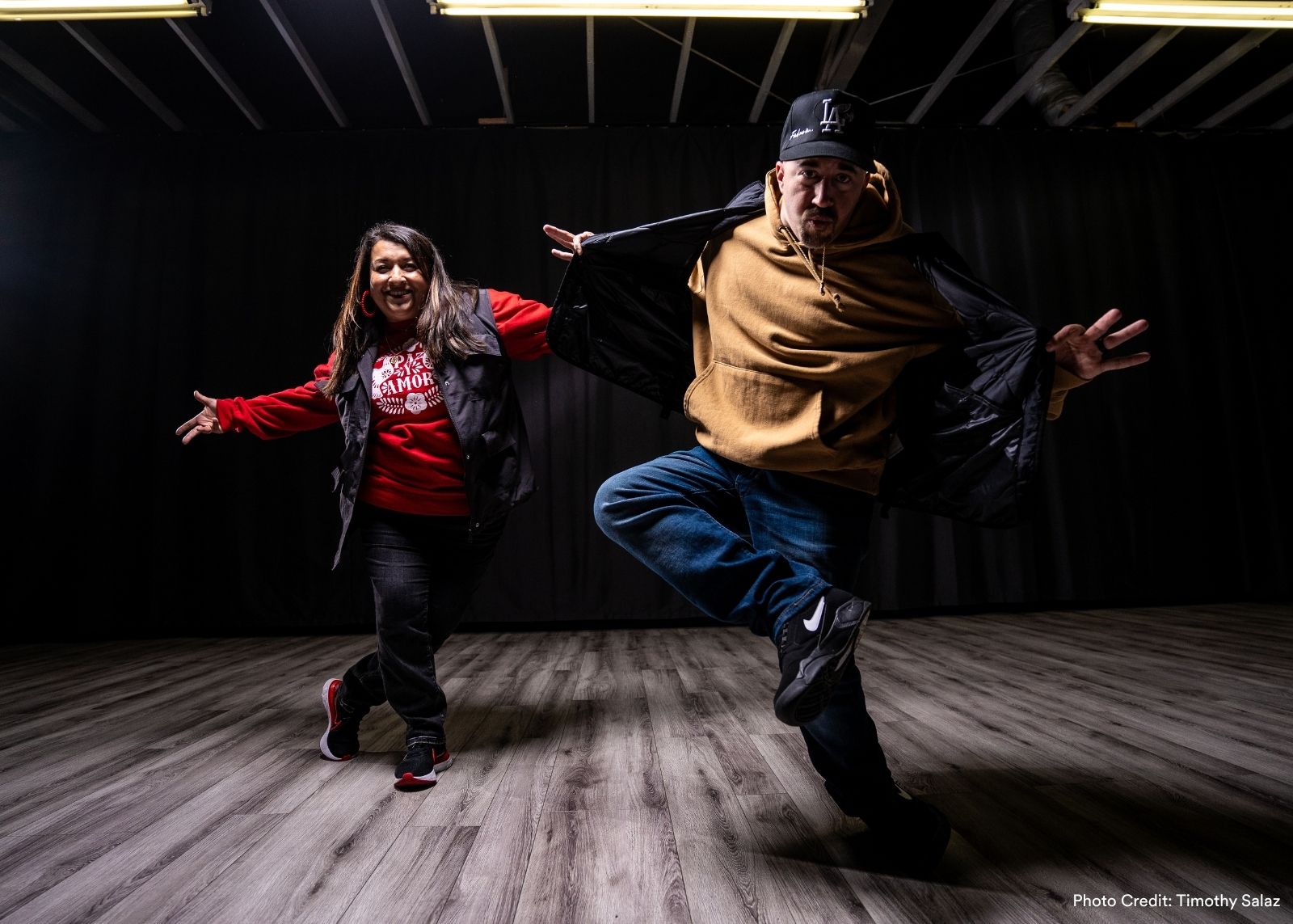 Two Versa-Style dancers perform in a studio. One leaps mid-air with jacket open while the other smiles with arms out. Photo: Timothy Salaz.