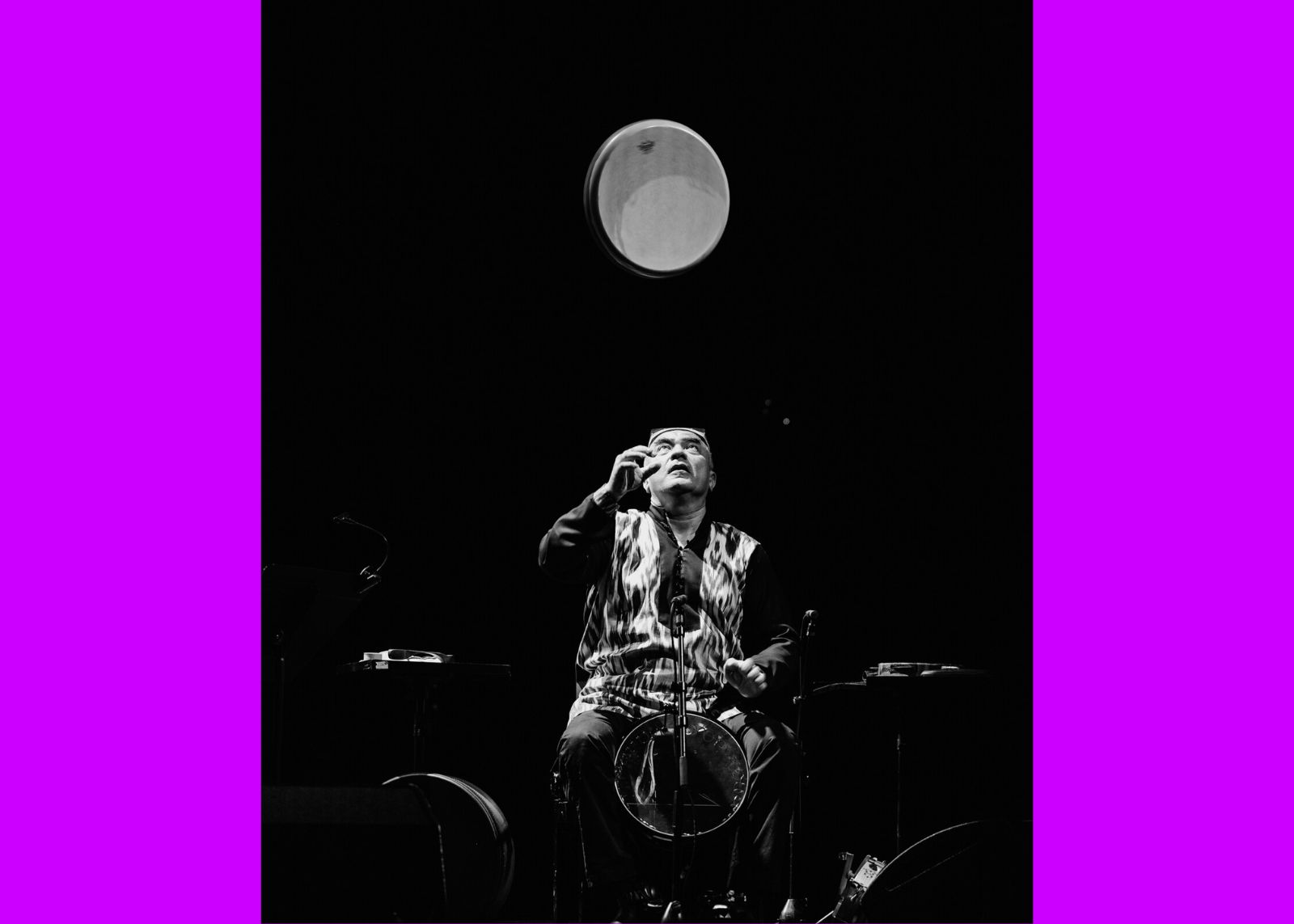 A black-and-white photo of a musician tossing a frame drum into the air on a darkened stage.
