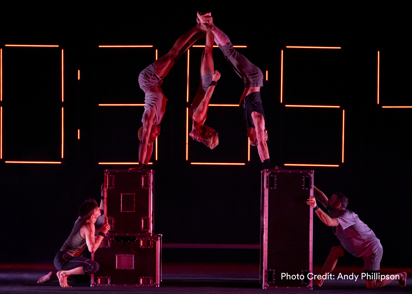 Two acrobats form an arch with their bodies atop road cases, hands clasped above, as two others brace the cases below. (Photo Credit: Andy Phillipson)
