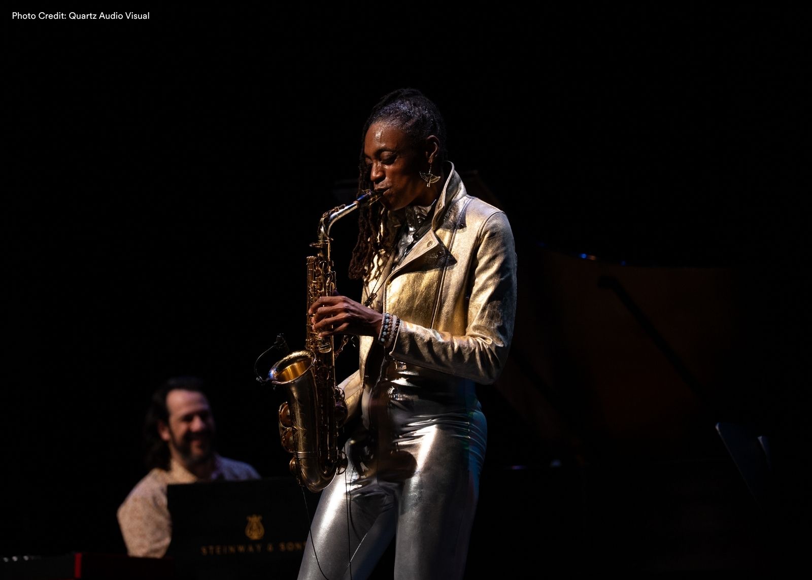 A saxophonist in a gold metallic jacket and silver pants performs on a darkened stage, with a pianist visible and a Steinway grand piano in the background.