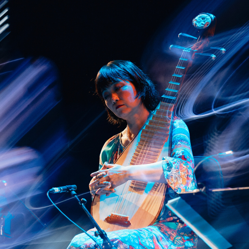 A pipa player sits in front of a black backdrop with light swirling around her.