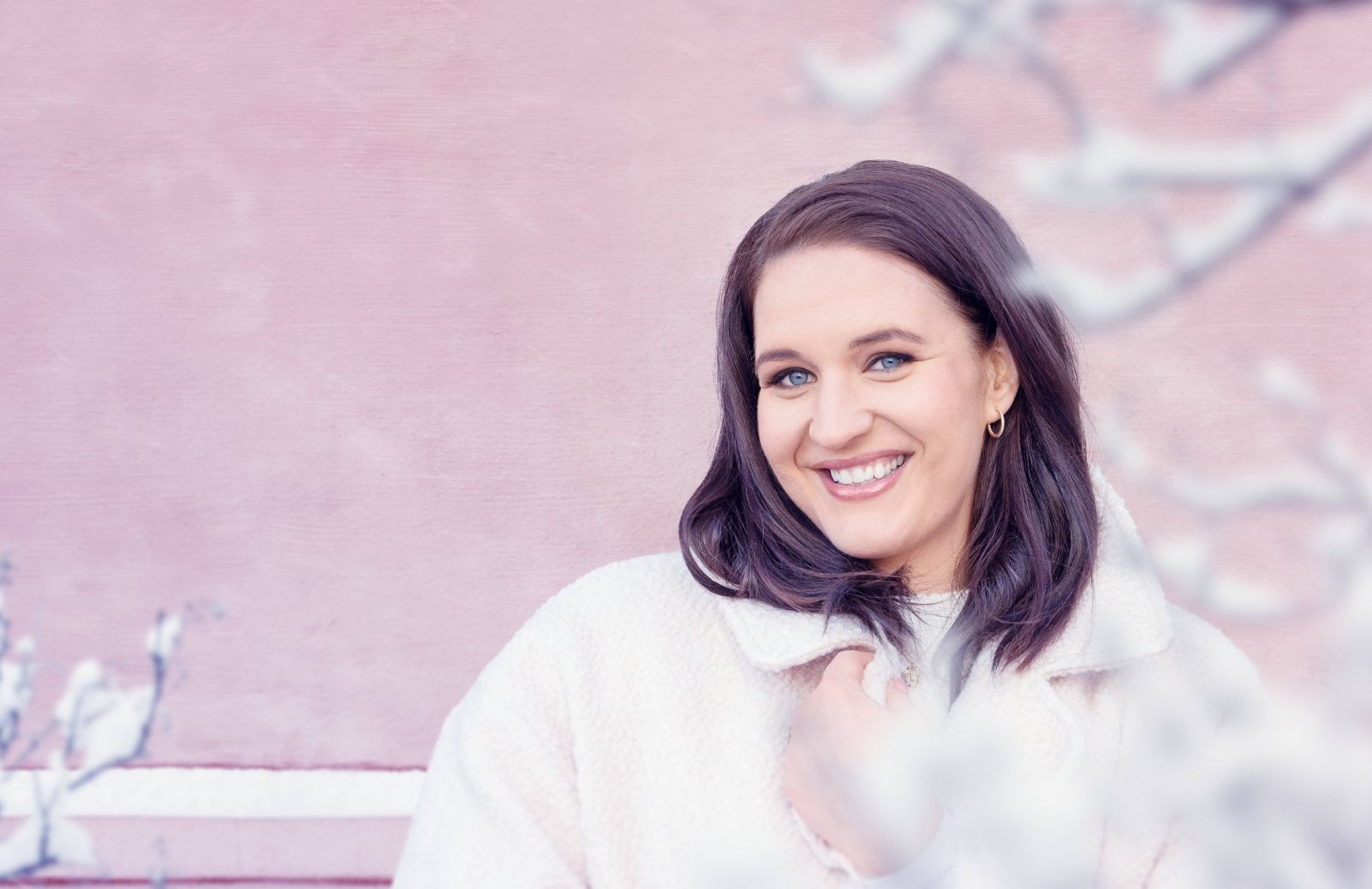 A woman in a white coat smiles, in front of a pink wall with white branches in the foreground.