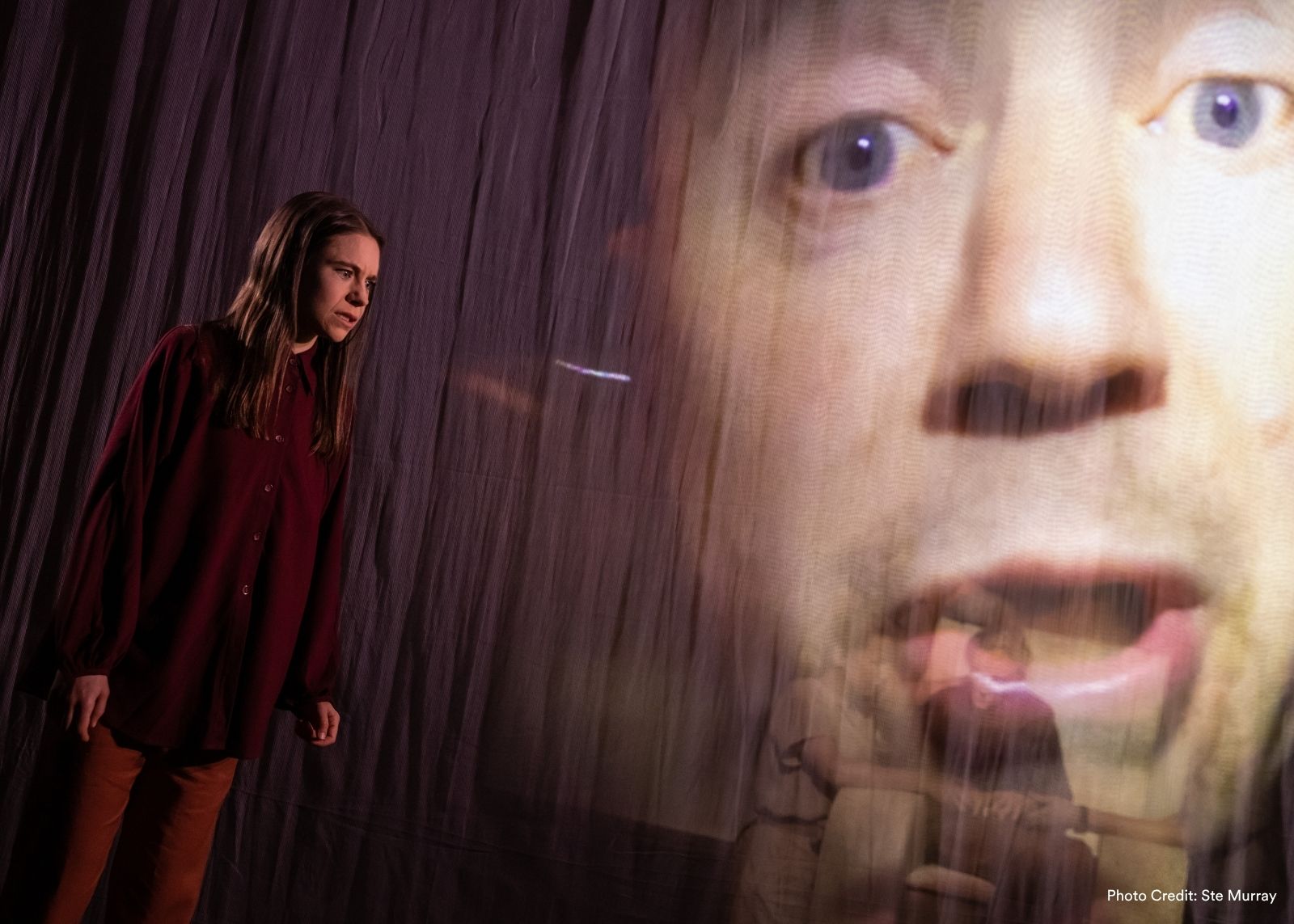 A performer looks tense beside a large projected close-up of a face on a draped stage backdrop.