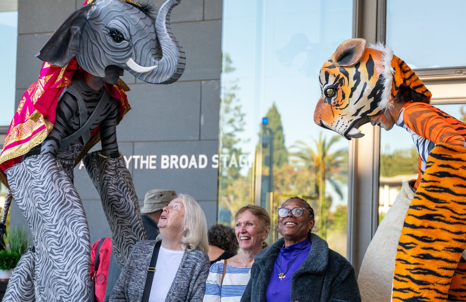 Three audience members laugh and look up at elaborate elephant and tiger puppets outside the BroadStage entrance.