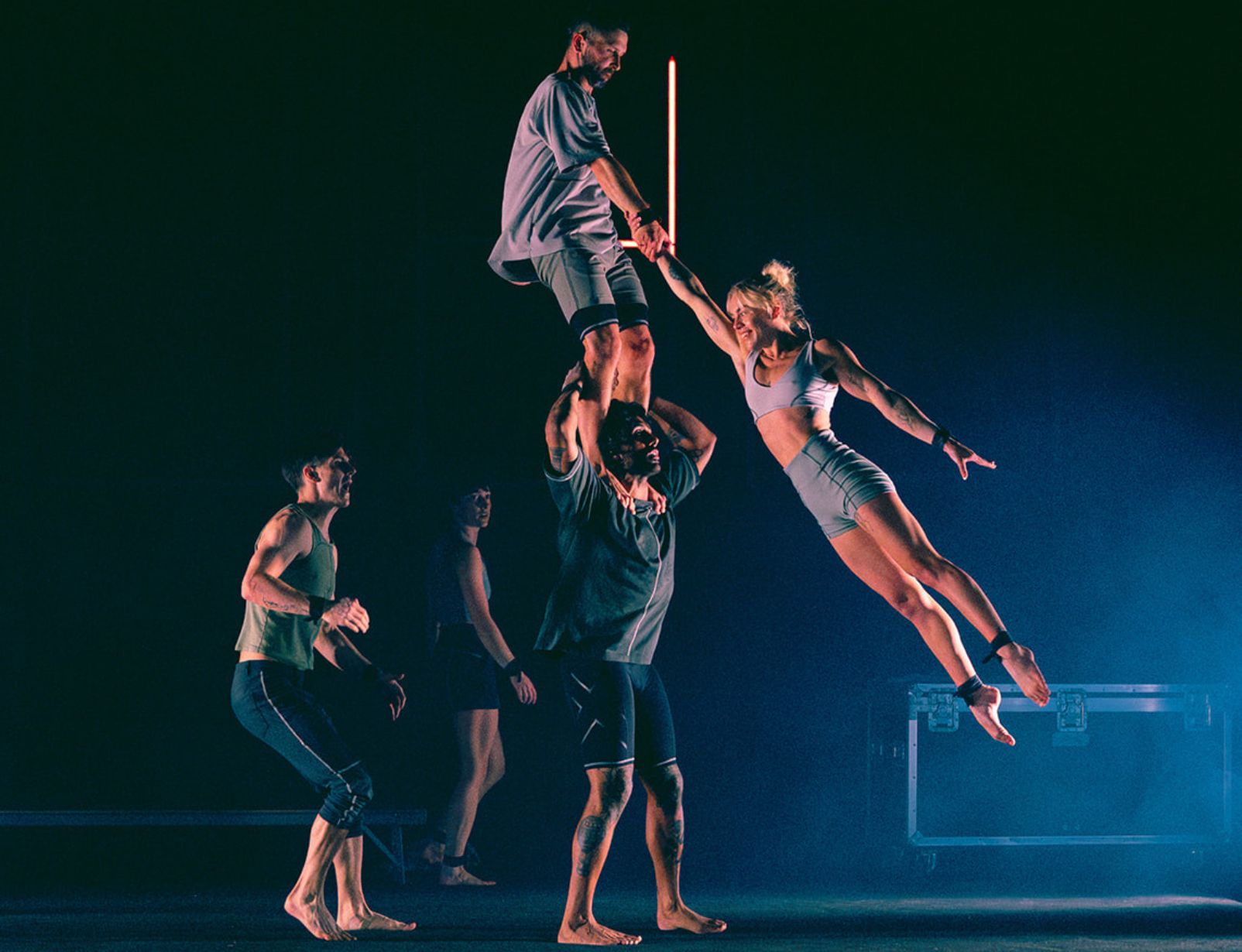 Acrobatic performers form a human tower on a dark stage, one dancer extended horizontally in the air.