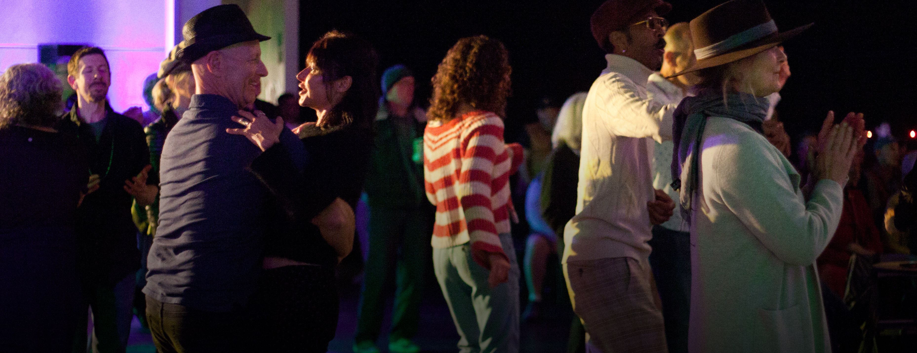 Audience members dance together at an outdoor evening event under colorful stage lighting.