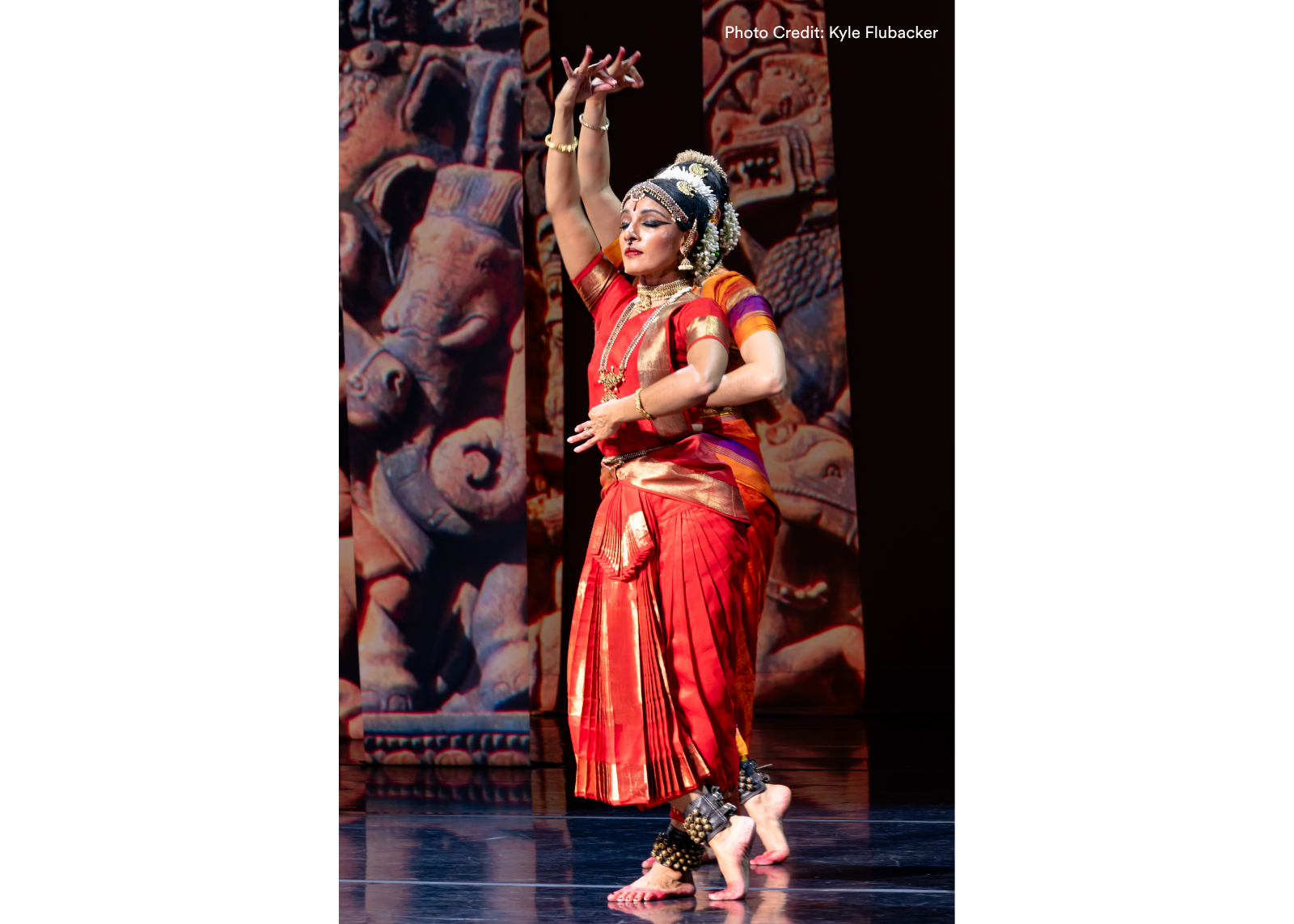 Two dancers in red and gold silk costumes and ankle bells perform with one arm raised, against a backdrop of projected stone carvings. Photo: Kyle Flubacker.