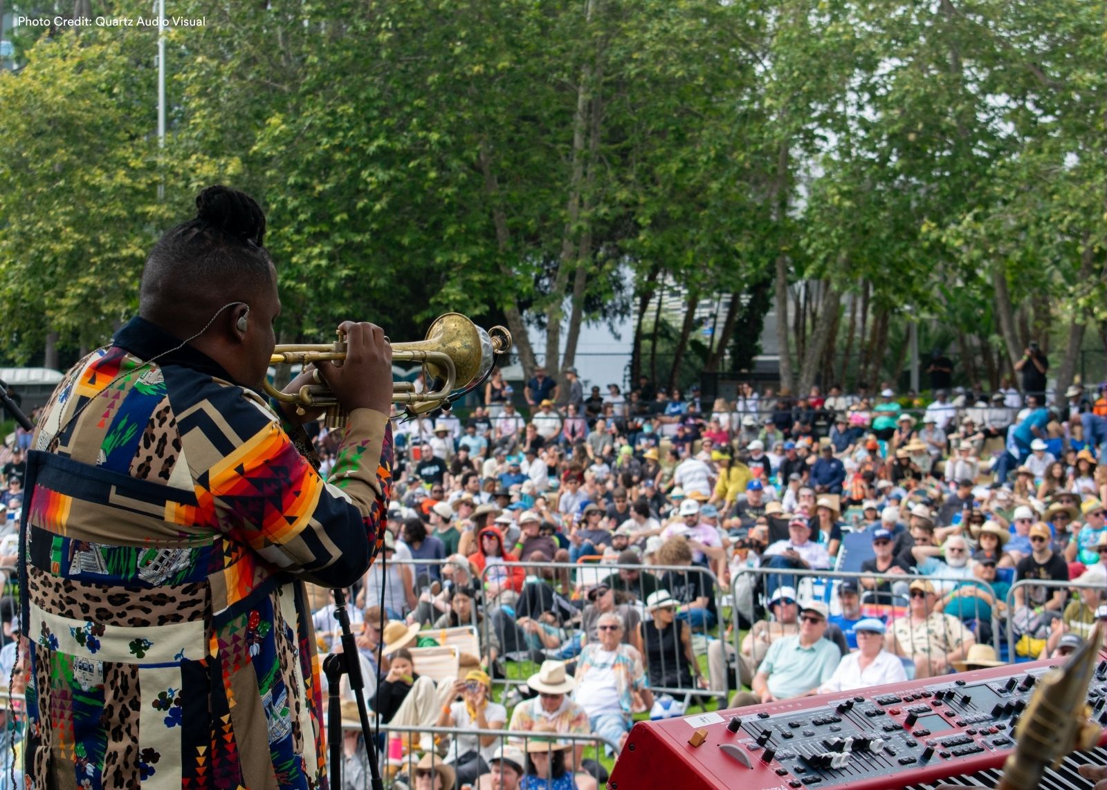 A trumpeter in a bold patchwork coat performs with their back to the camera before a large outdoor festival crowd.