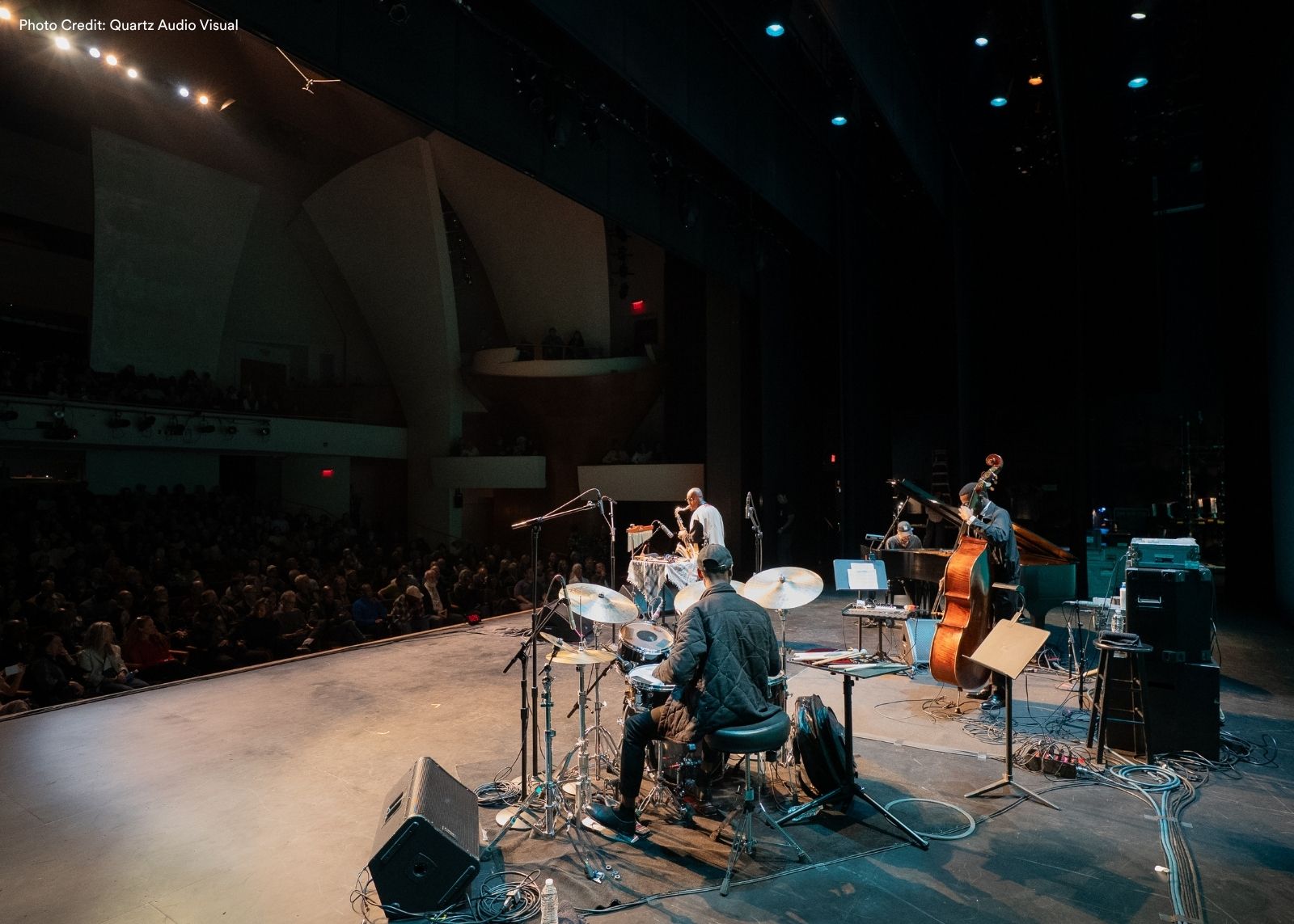 A wide shot of a jazz quartet performing on a concert stage to a packed house, with a drummer in the foreground and a double bassist, pianist, and saxophonist visible behind.