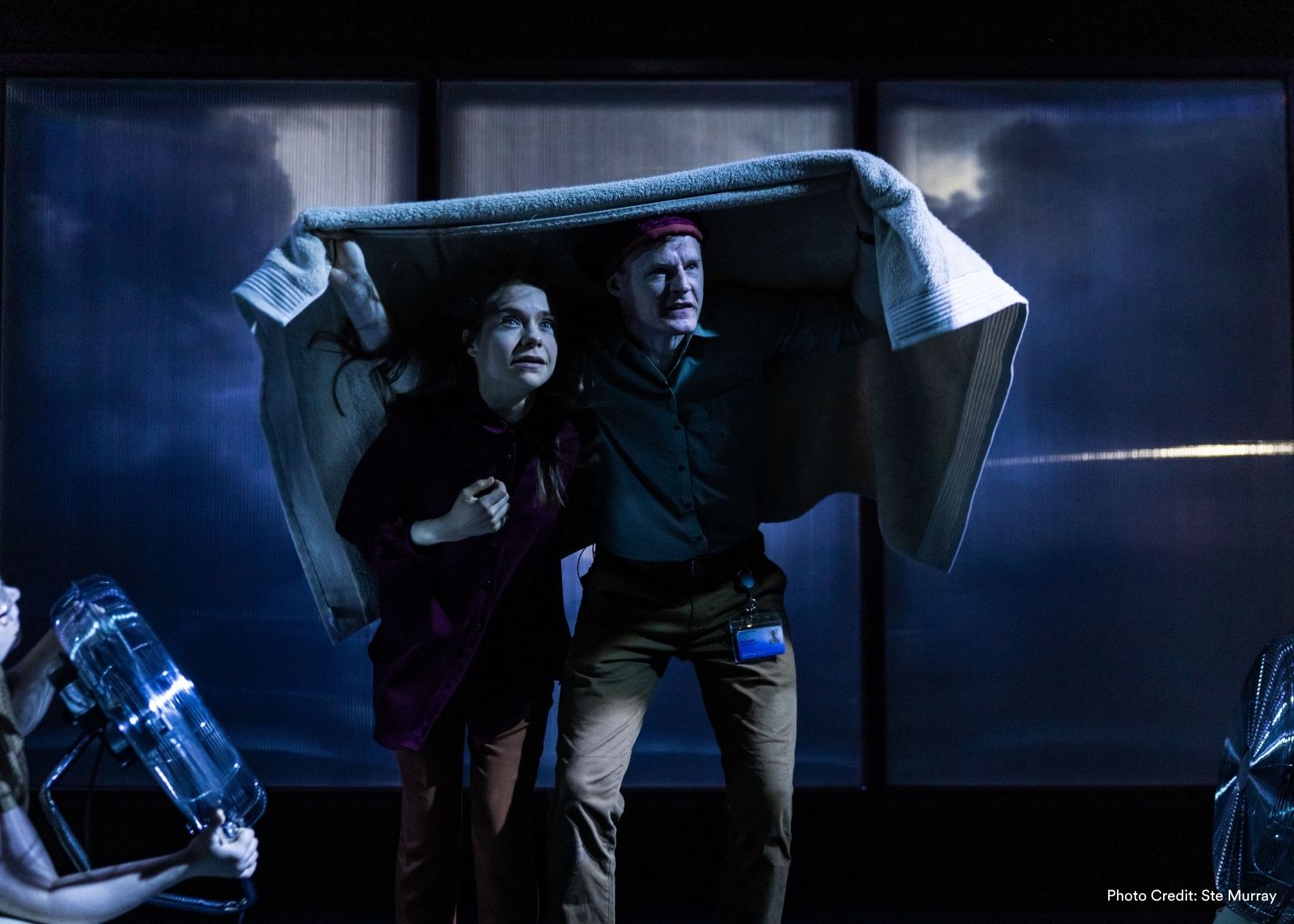Two performers shelter under a blanket on a stormy blue-lit stage, flanked by cast members holding electric fans.